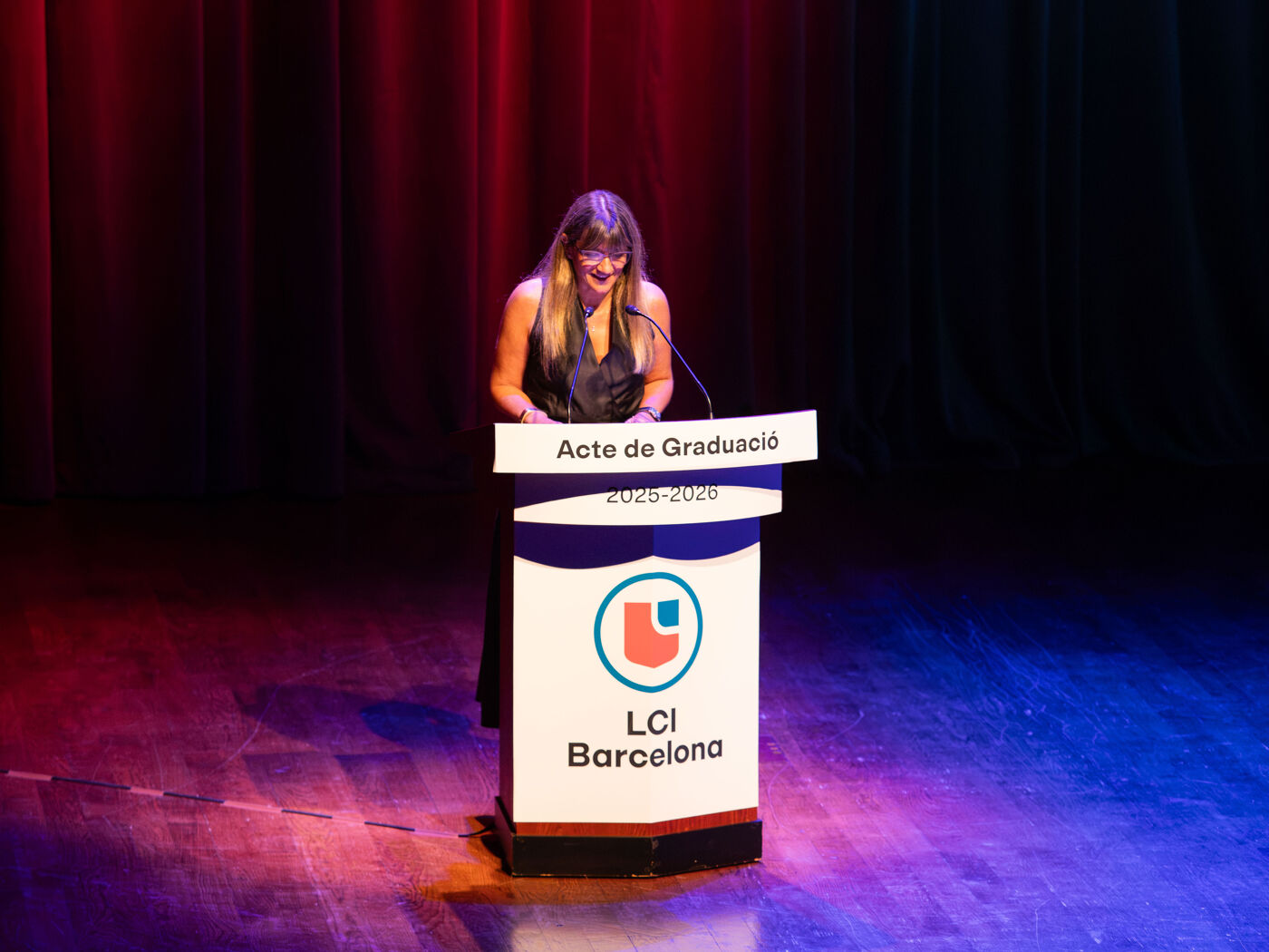 A woman speaks from a podium with the LCI Barcelona logo, during a graduation ceremony.