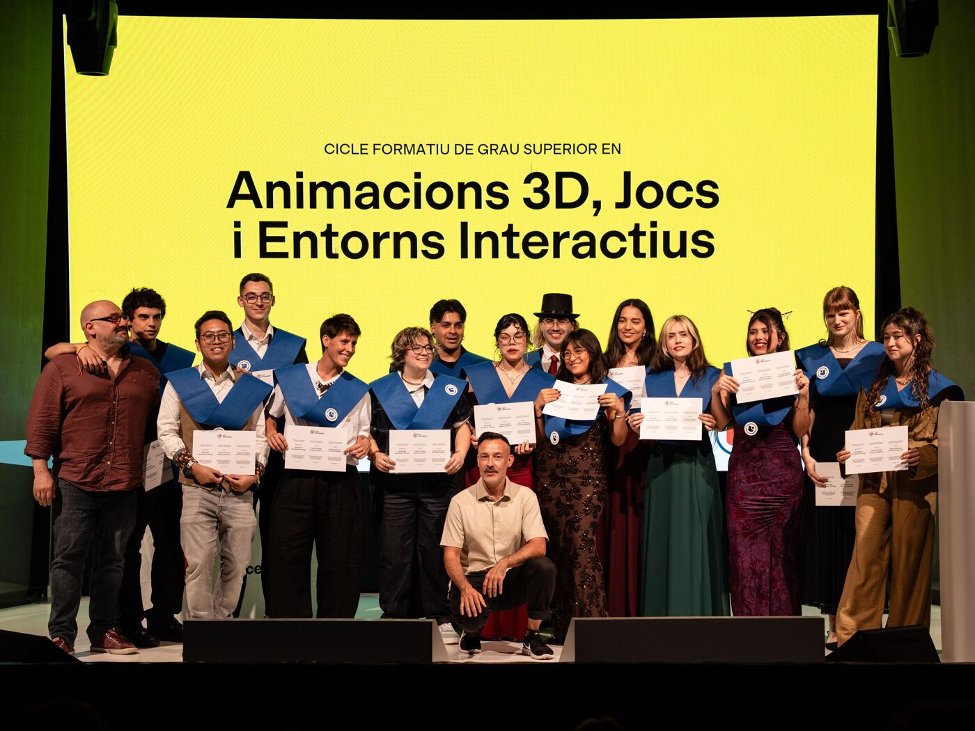 A group of graduates pose for a photo with their certificates, with a large yellow banner behind them reading "3D Animations, Games and Interactive Environments".