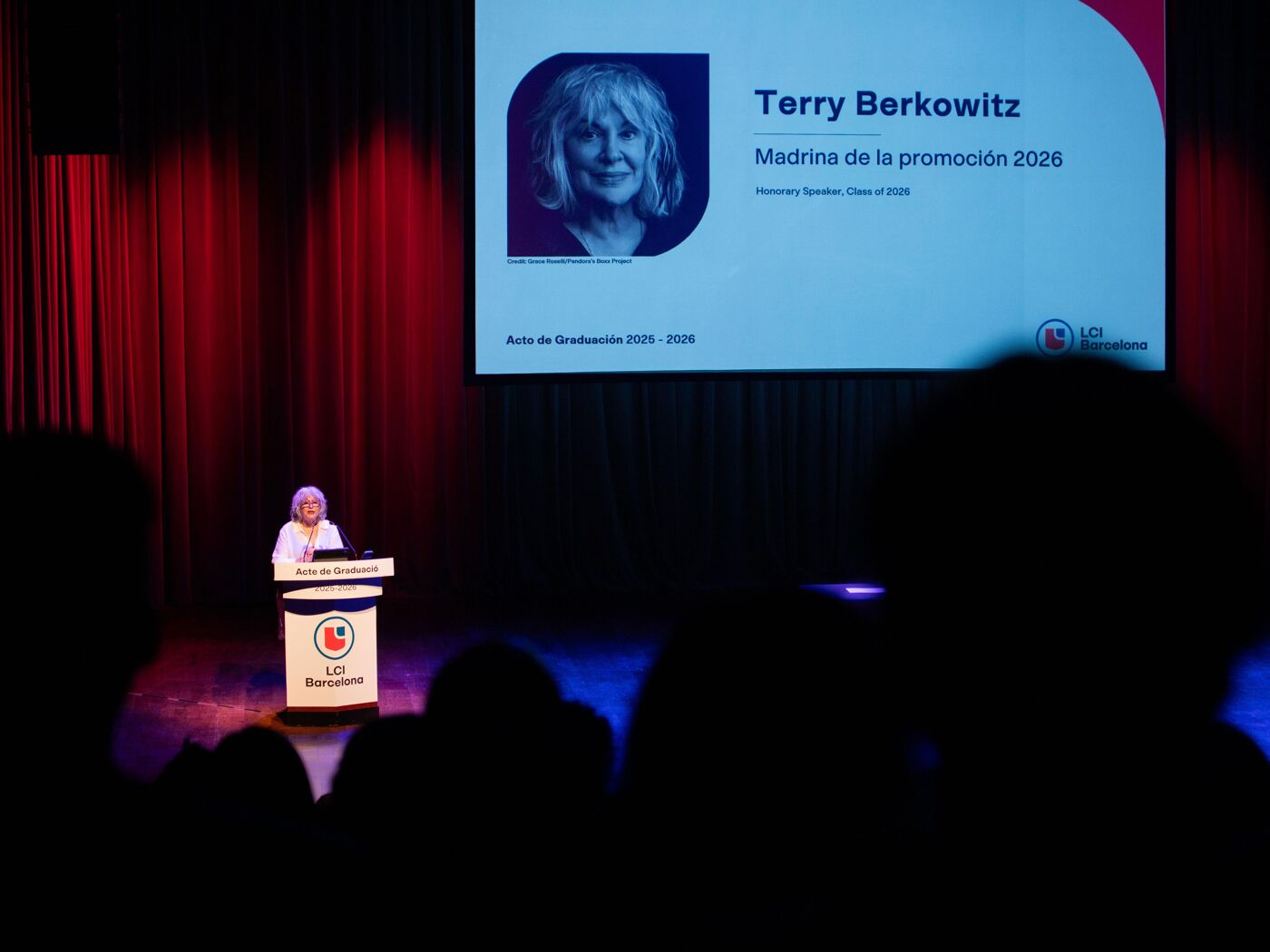A woman speaks from a podium on a stage in front of an audience. Behind her, a screen displays a photo of Terry Berkowitz and the text "Madrina de la promoción 2026".