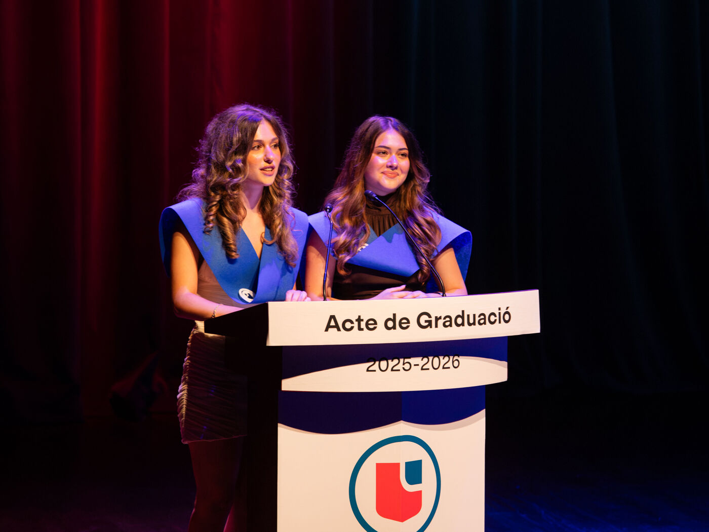 Two young women in blue academic regalia stand behind a podium that reads "Acte de Graduació 2025-2026". They appear to be addressing an audience during a graduation ceremony.