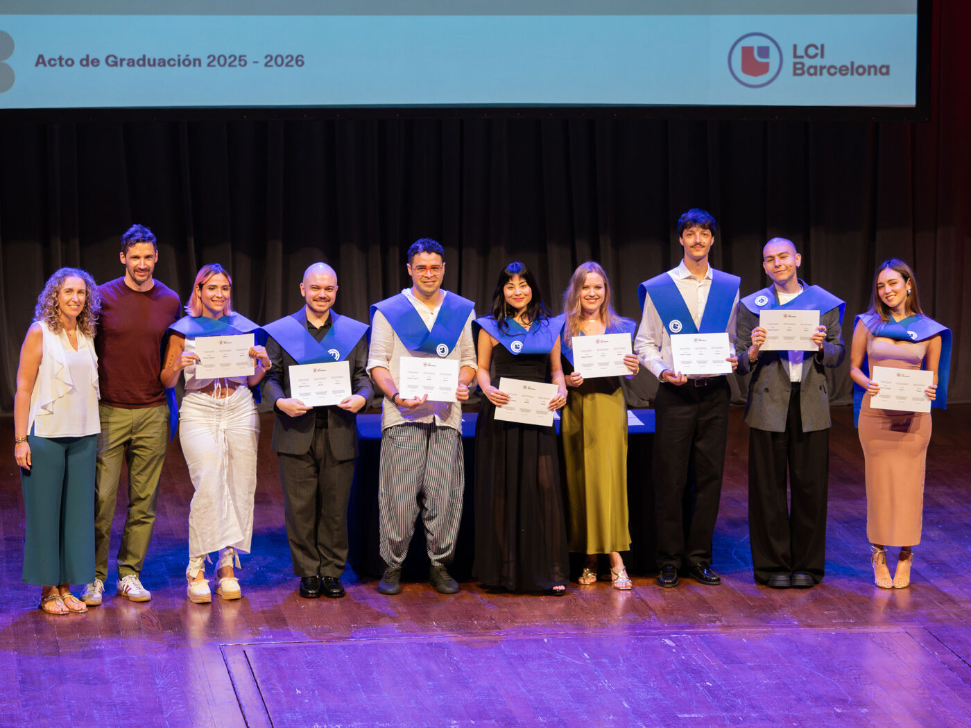 A group of graduates pose for a photo holding their certificates, dressed in blue and white academic regalia.