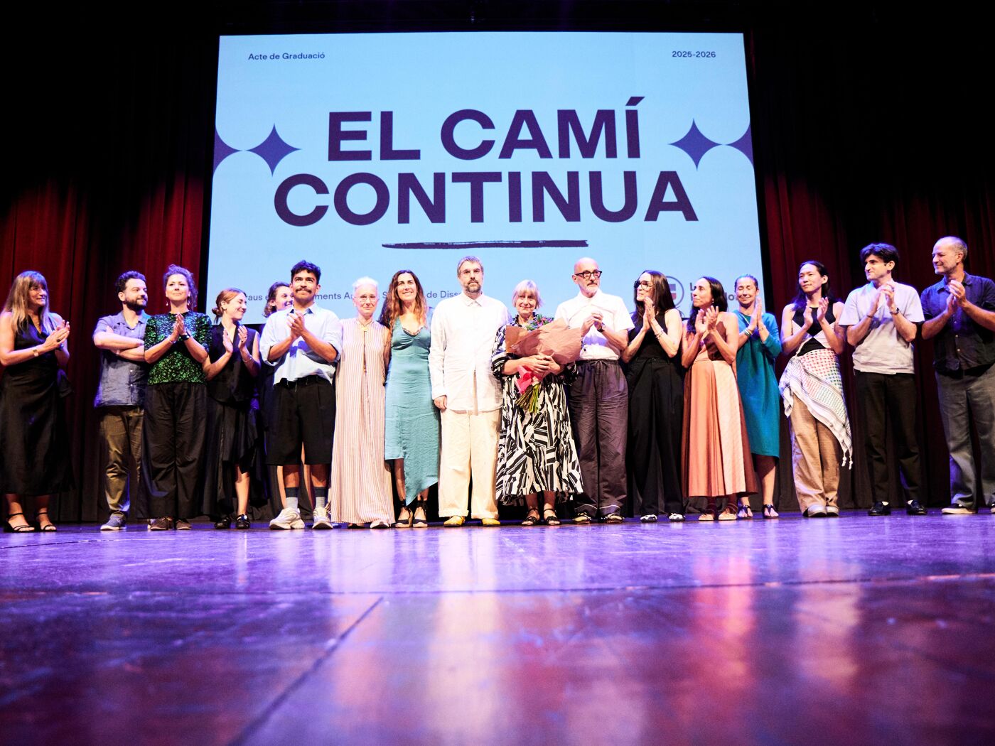 A group of people stand on a stage under a large banner that reads 'EL CAMÍ CONTINUA'. It appears to be a celebration or a graduation event.