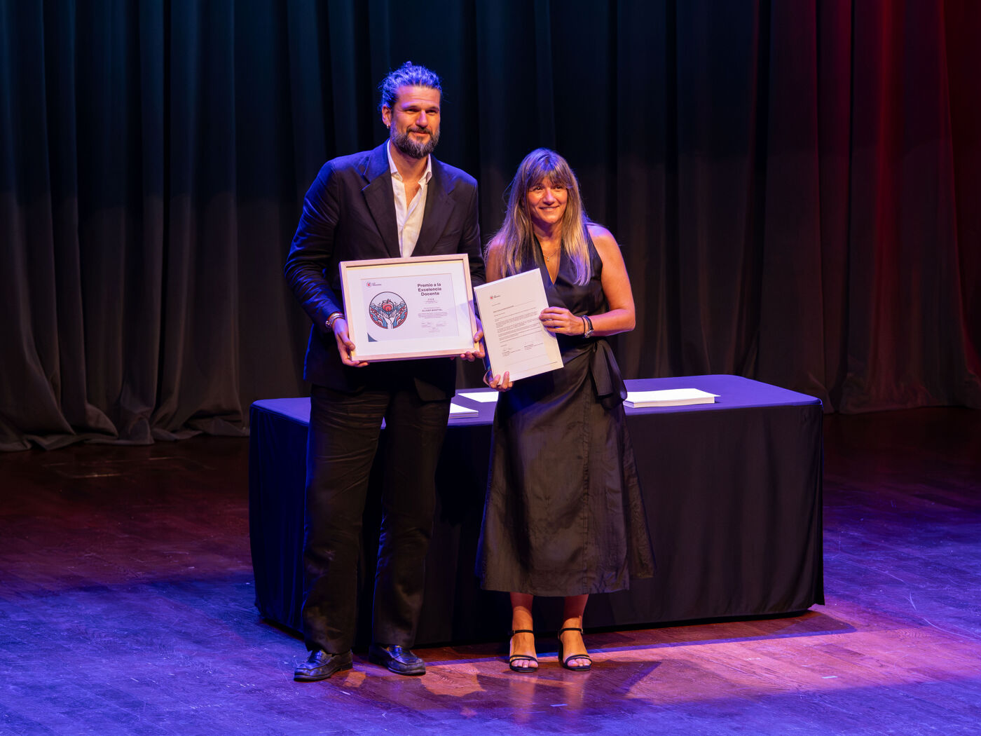 Two people are on a stage receiving awards. The man is holding a framed certificate, and the woman holds a document in her hand.