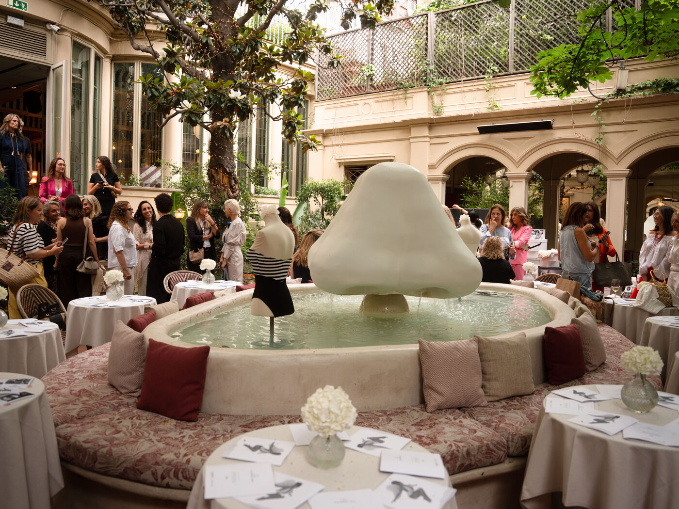 A mushroom-shaped fountain with circular seating surrounds it, with tables and chairs arranged around in an indoor courtyard with trees and classical-style architecture.