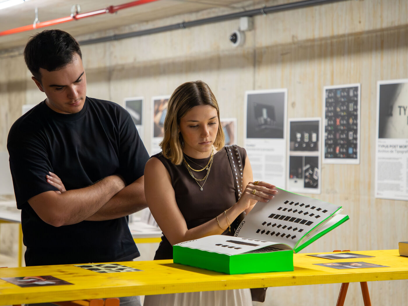 Two people review an open design portfolio with pattern samples at a yellow table in an indoor gallery or studio.
