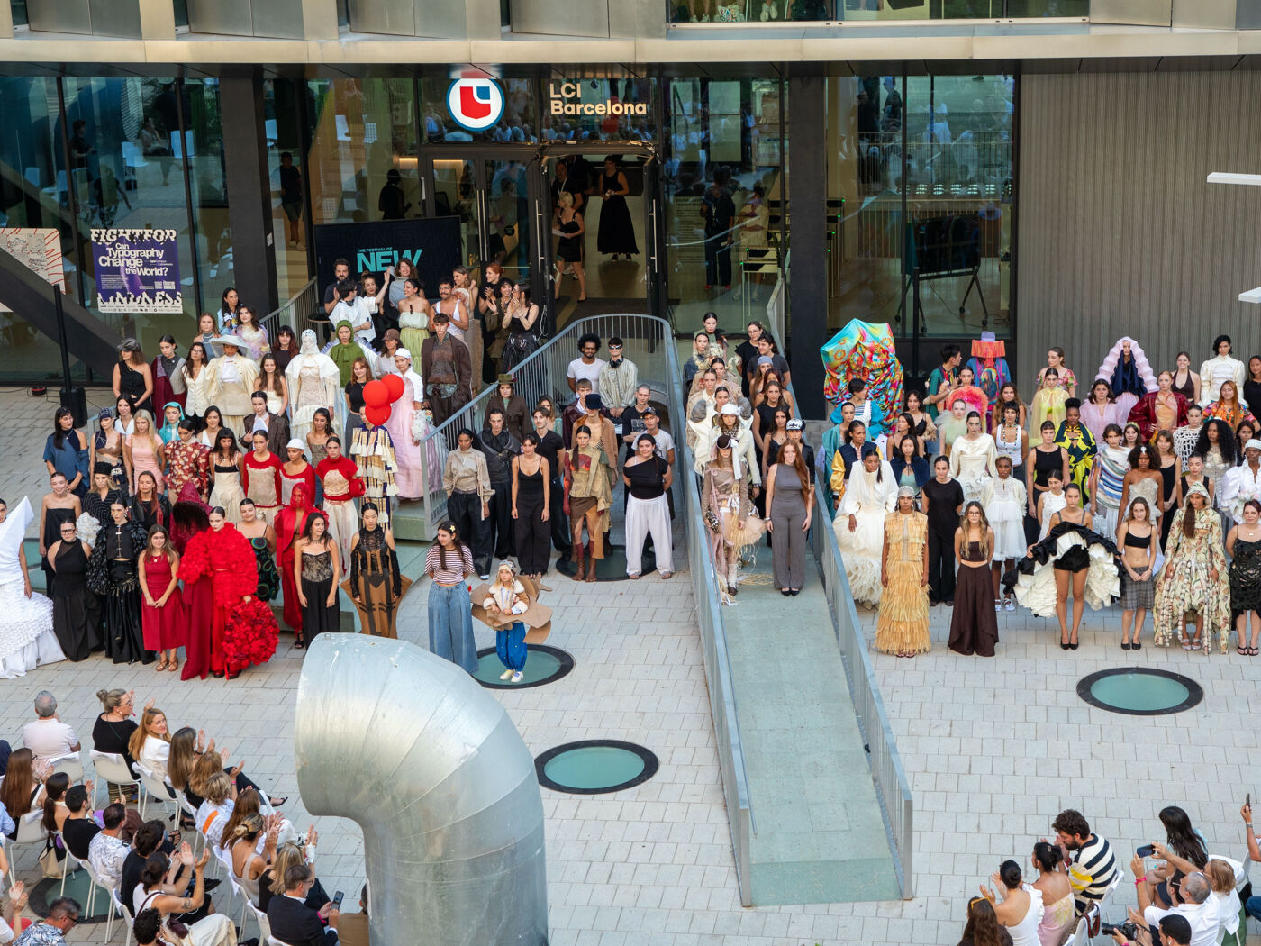 Wide outdoor group photo of models in varied runway looks posing at the entrance of LCI Barcelona during NEW'26 – The Festival of New Ideas, with a seated audience in the foreground.