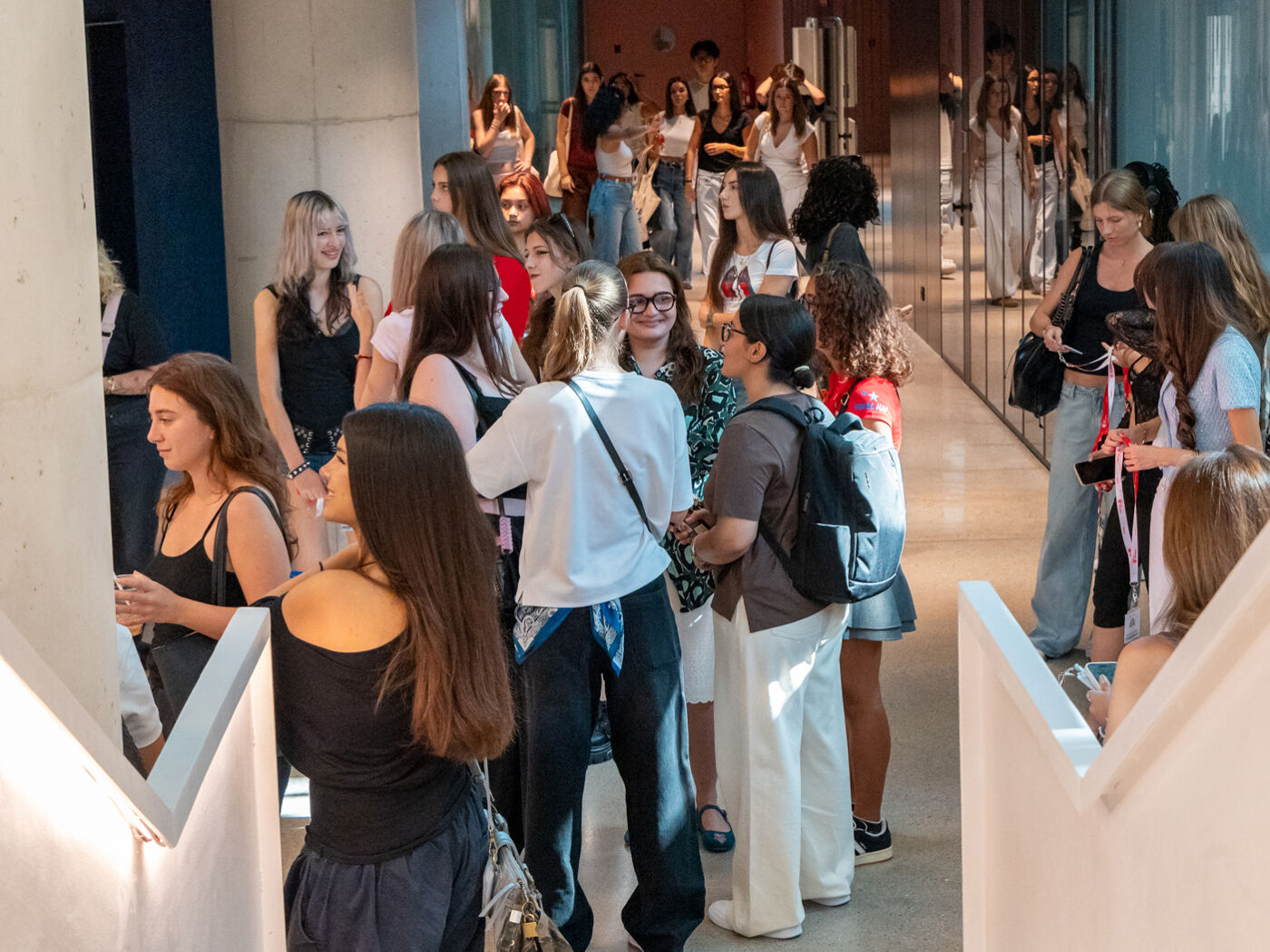A group of young women are gathered in a long, bright hallway, some talking amongst themselves while others look towards a point out of view.