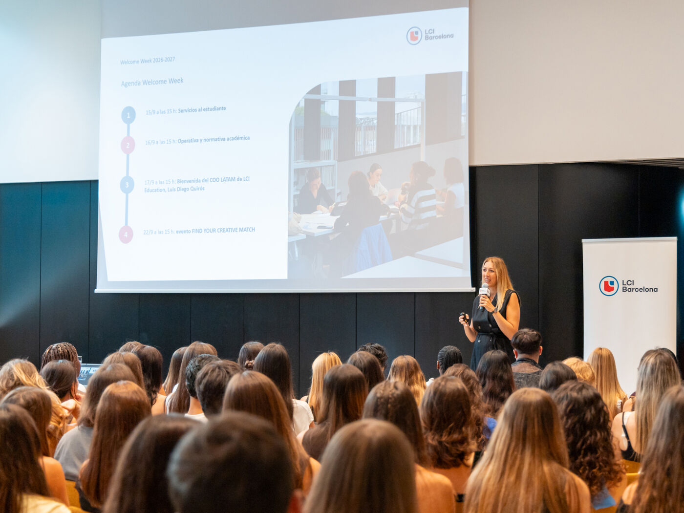 A woman is speaking to a large audience in a conference room, with a presentation displayed on a screen behind her.