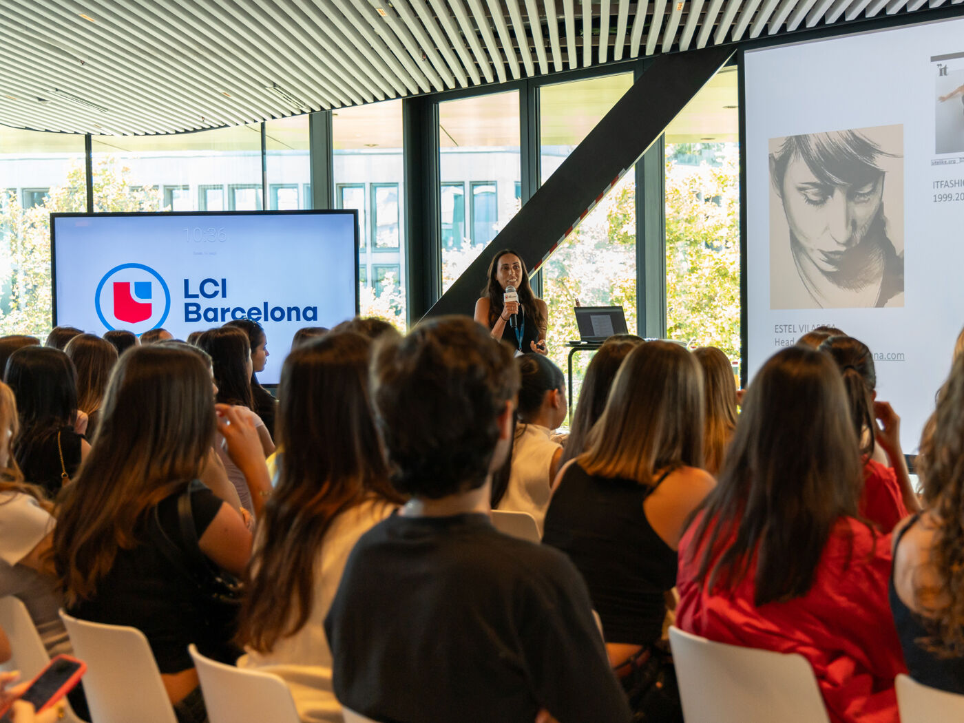 A woman speaks to an audience seated in an event room in front of a screen displaying the LCI Barcelona logo.