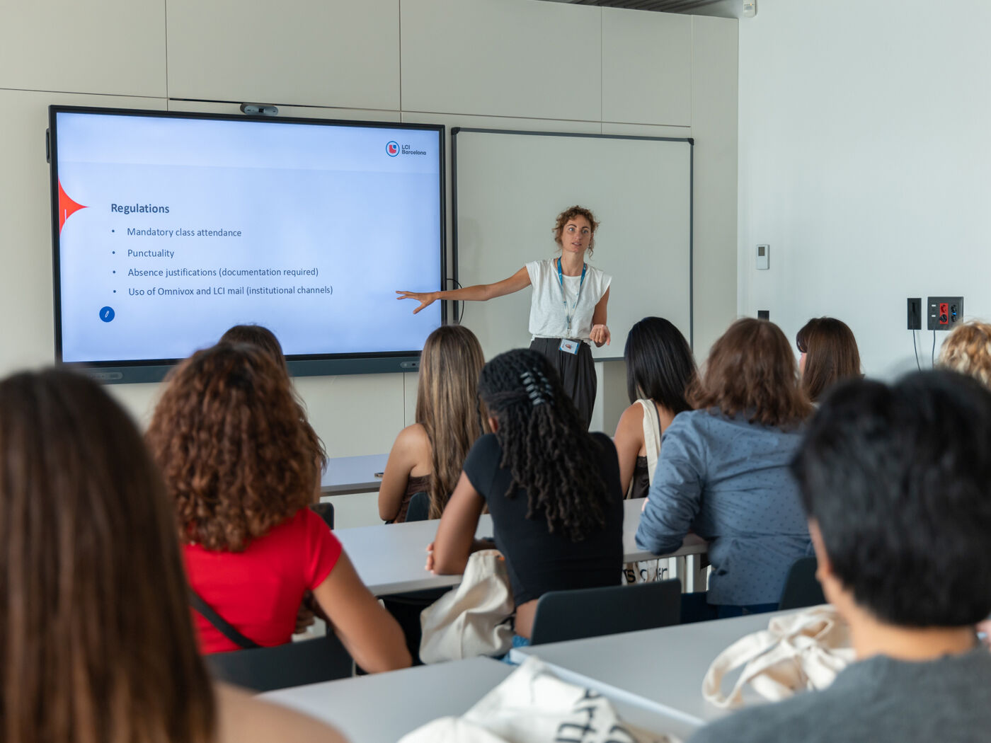 A woman is giving a lecture to a class of students, pointing to a presentation about regulations on a large screen.