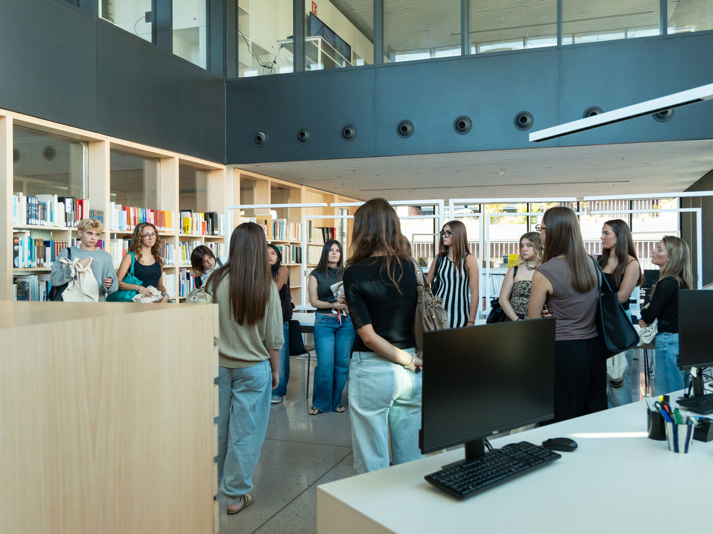 A group of young women are gathered in a modern library, attentively listening to a woman who appears to be giving a presentation or a guided tour.