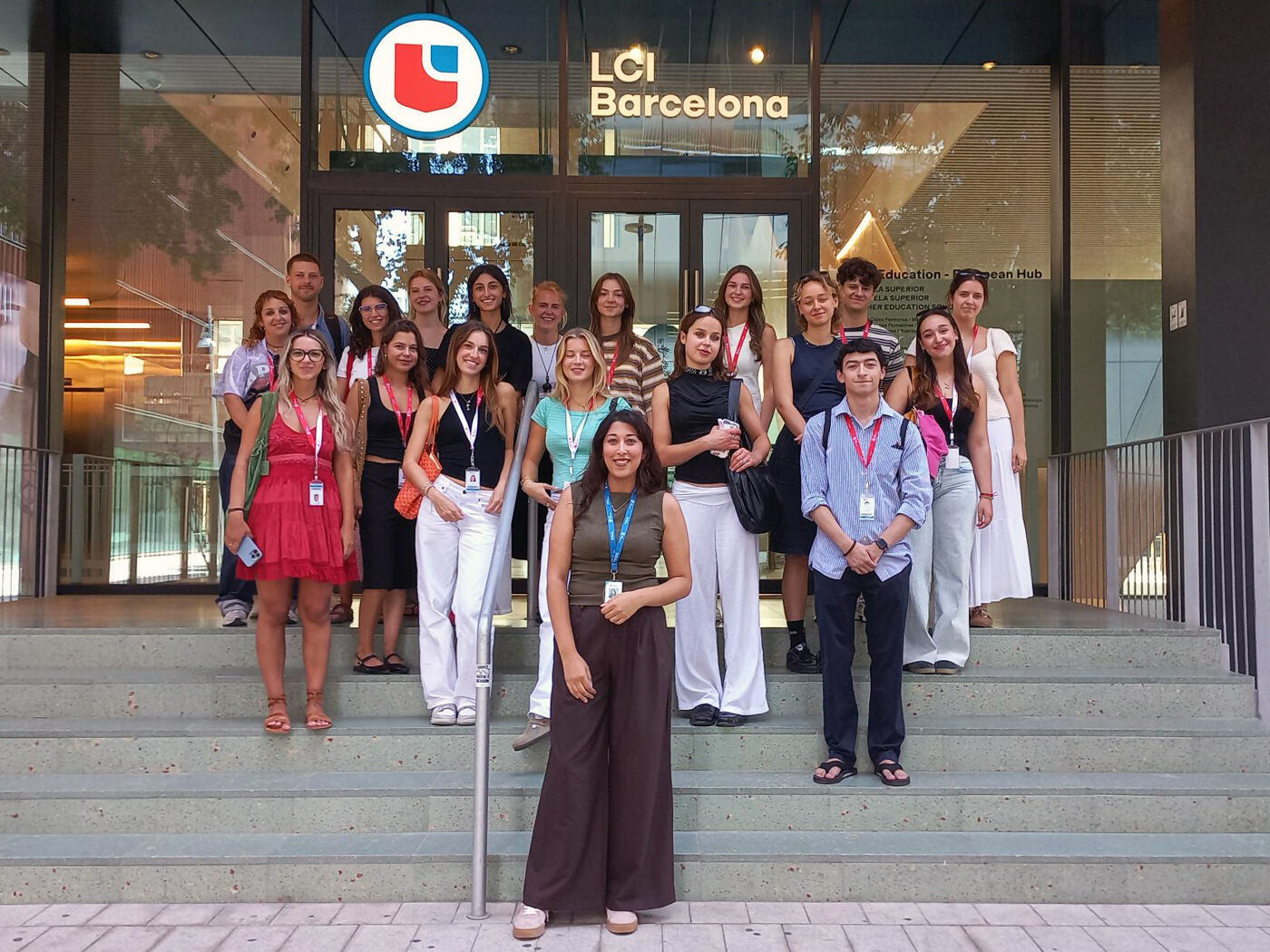 A group of people are gathered in front of the LCI Barcelona building, some on the stairs and others at the entrance, posing for a group photo.