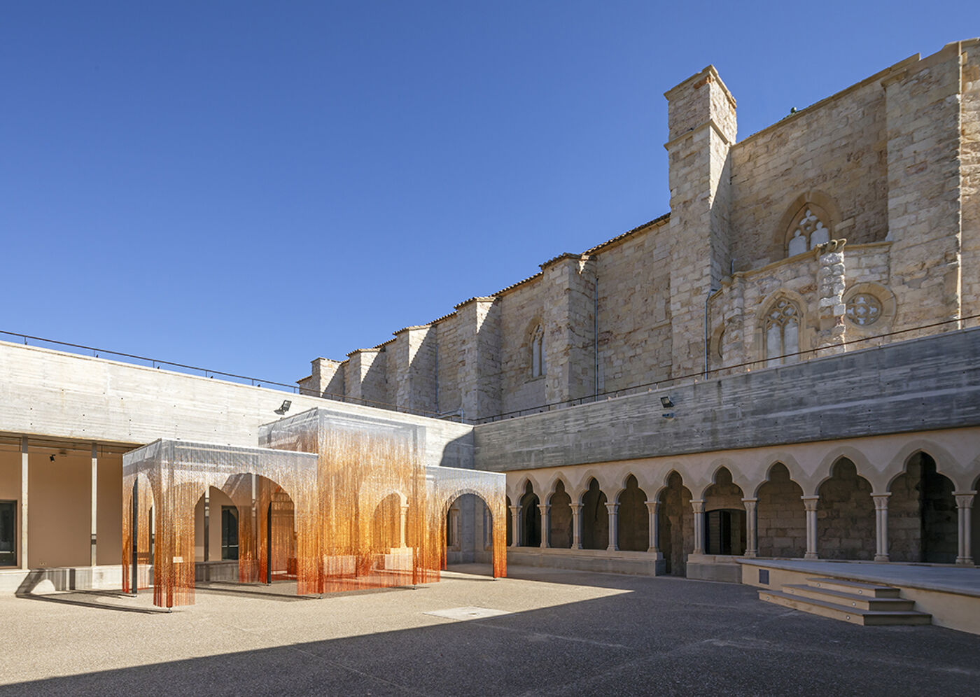 This image shows a modern, orange, translucent art installation set within the courtyard of a historic stone building with arched walkways and a clear blue sky above.