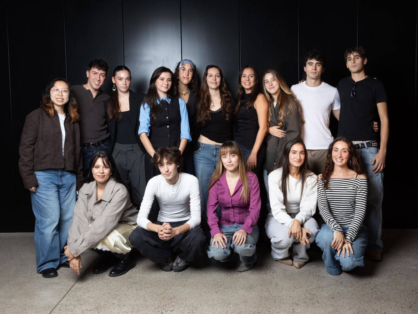 The image shows a group of 16 young adults, posing for a photo in front of a dark background. They are casually dressed and appear to be in their late teens or early twenties.