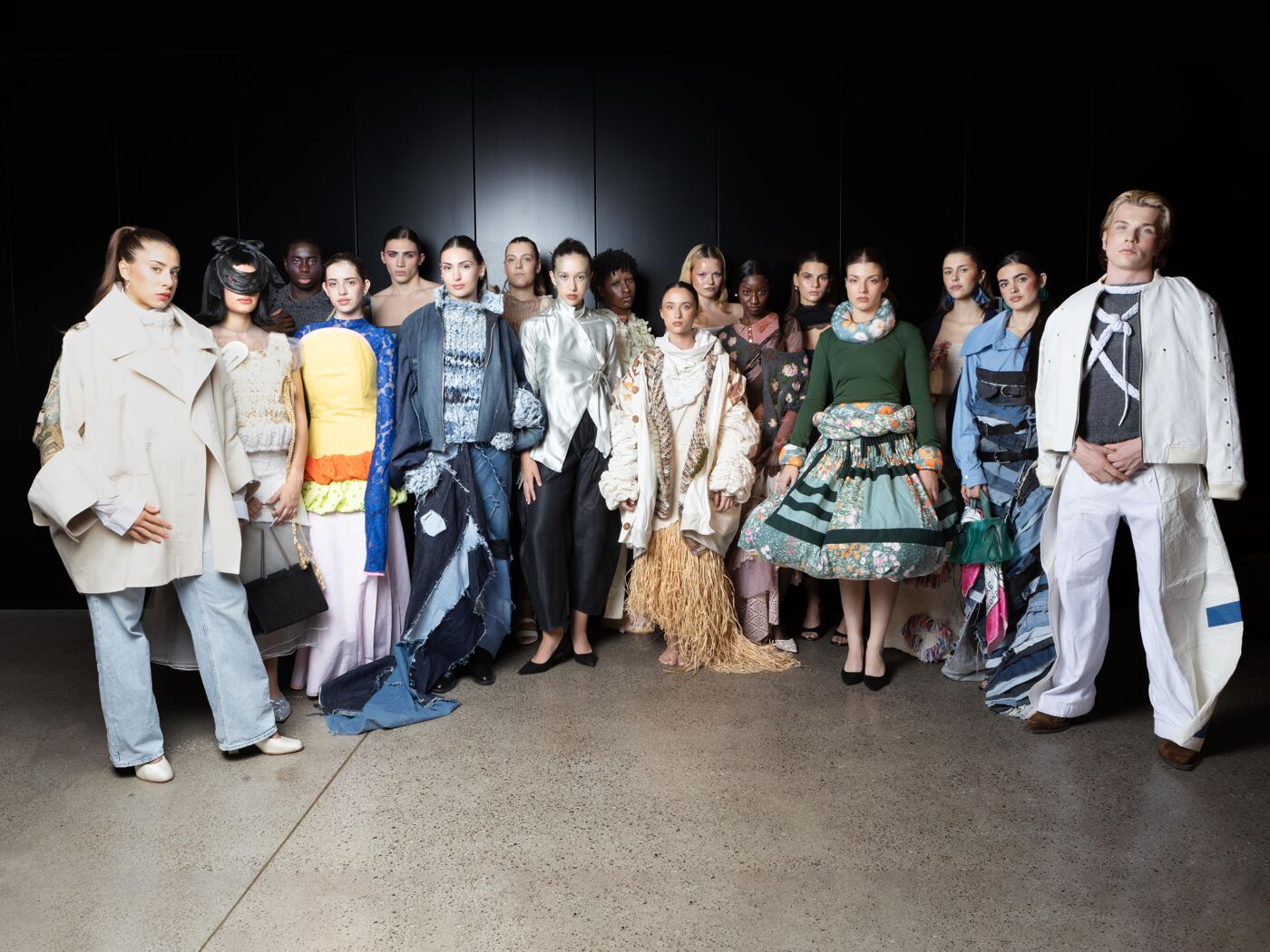 A diverse group of models, both male and female, pose together in a studio setting, showcasing avant-garde and deconstructed fashion pieces with a focus on denim and textured fabrics.