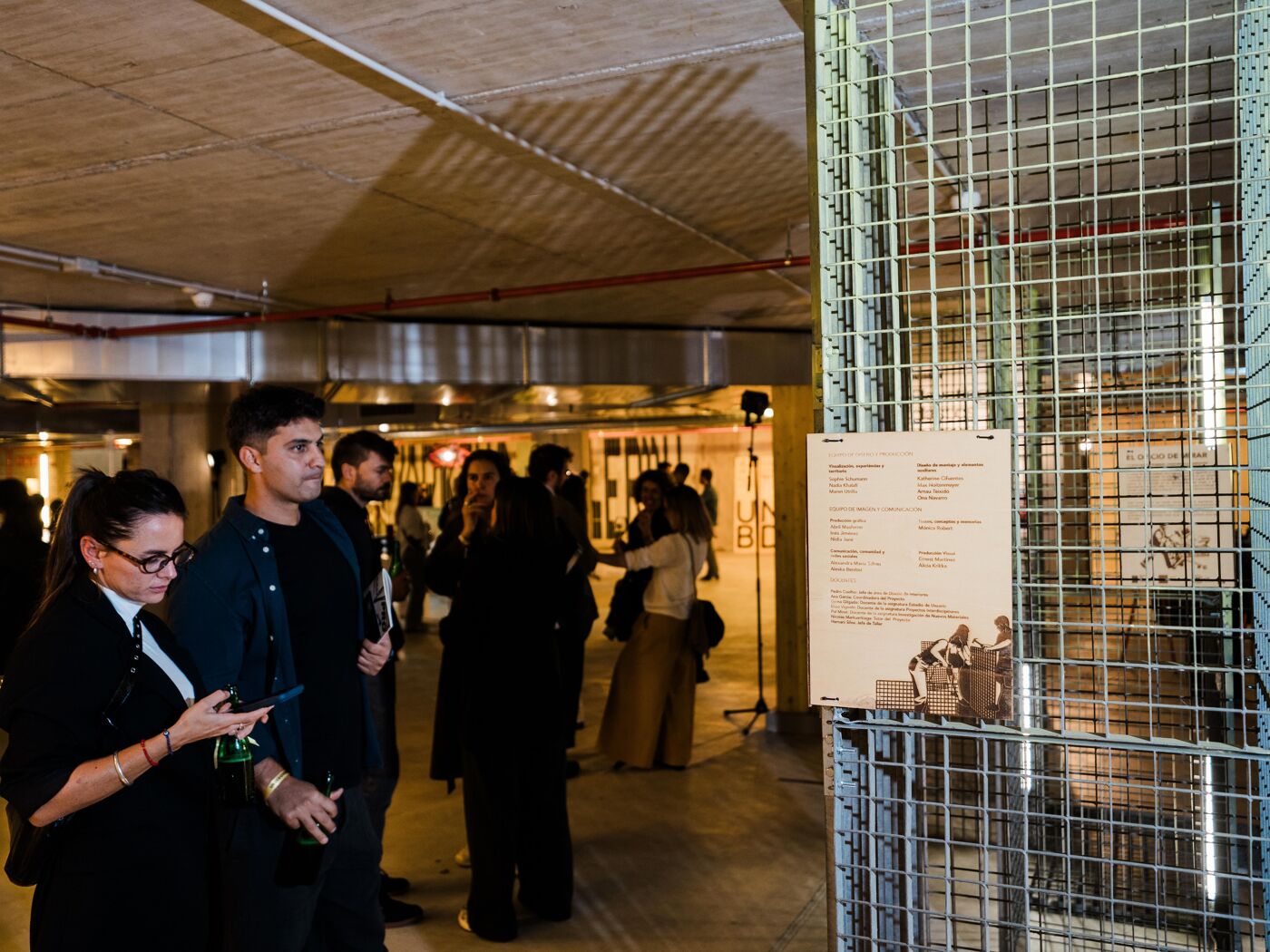 The image shows a group of people gathered indoors, possibly at an art exhibition or event. They are looking at displays and exhibits, with a focus on a metal grate structure with information panels.