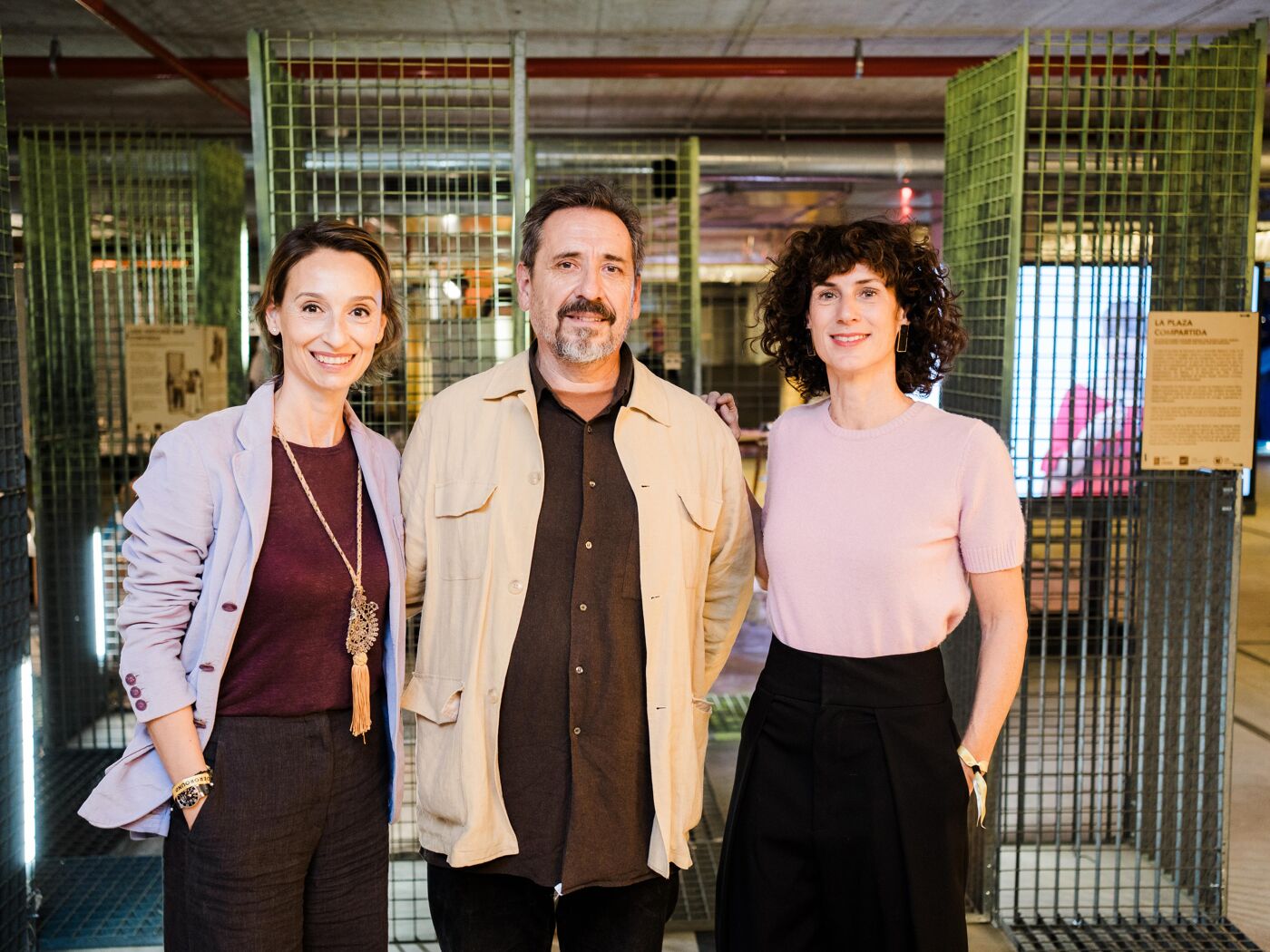 Three people, two women and one man, are standing in front of a metal cage-like structure. The woman on the left wears a purple blazer, the man in the middle wears a tan jacket, and the woman on the right wears a pink shirt.