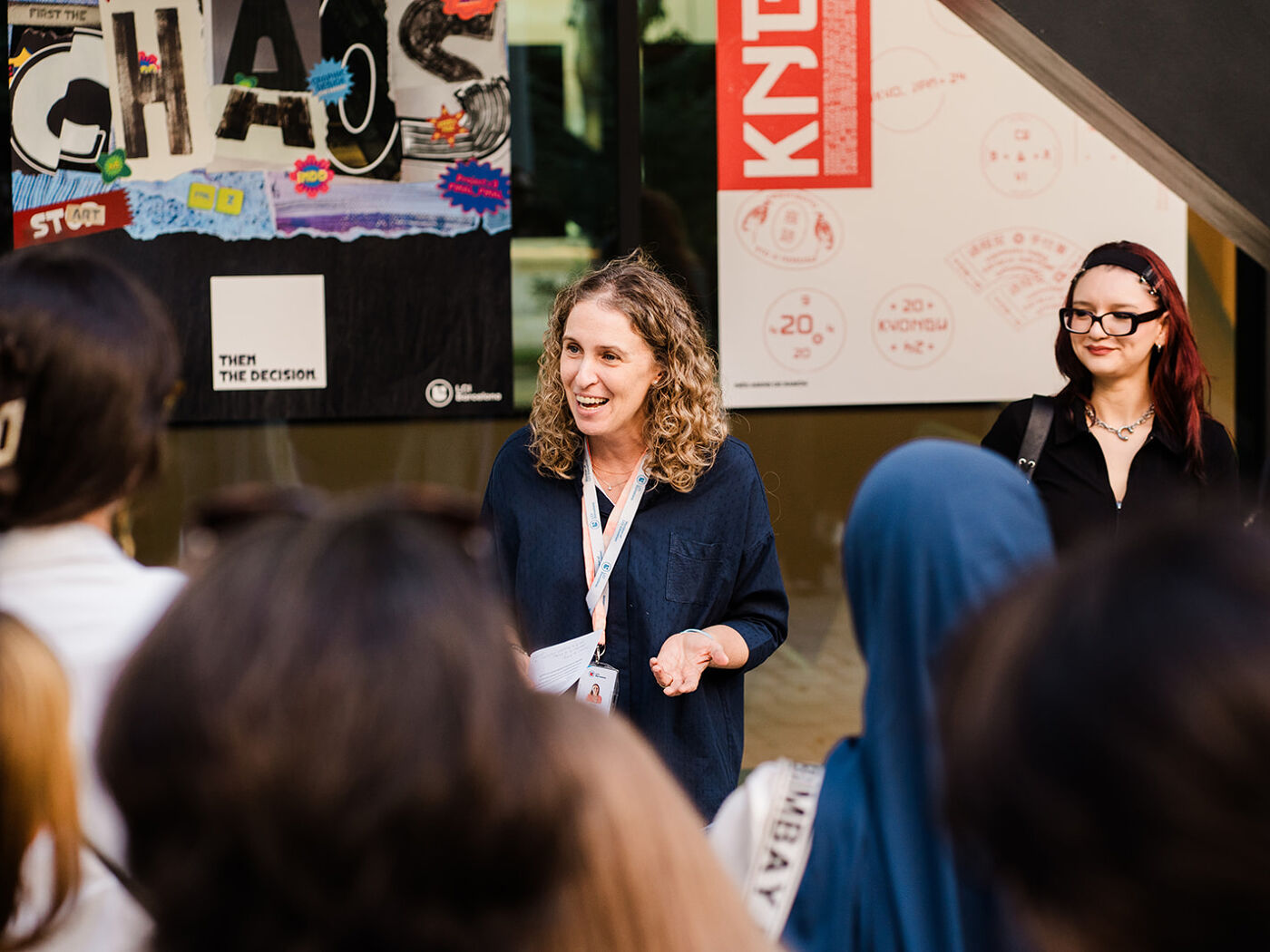A woman with curly hair is smiling and talking to a group of people, possibly giving a presentation or tour. Another woman stands behind her, observing.