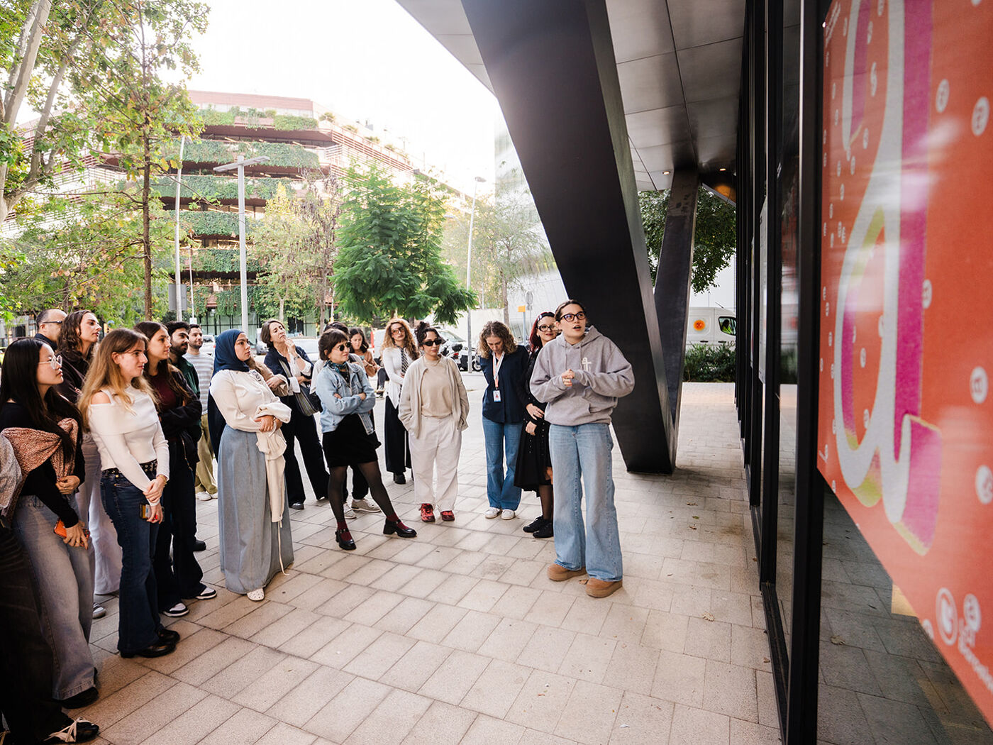 A group of people are gathered outside a modern building, listening to a woman who appears to be giving a presentation or tour.