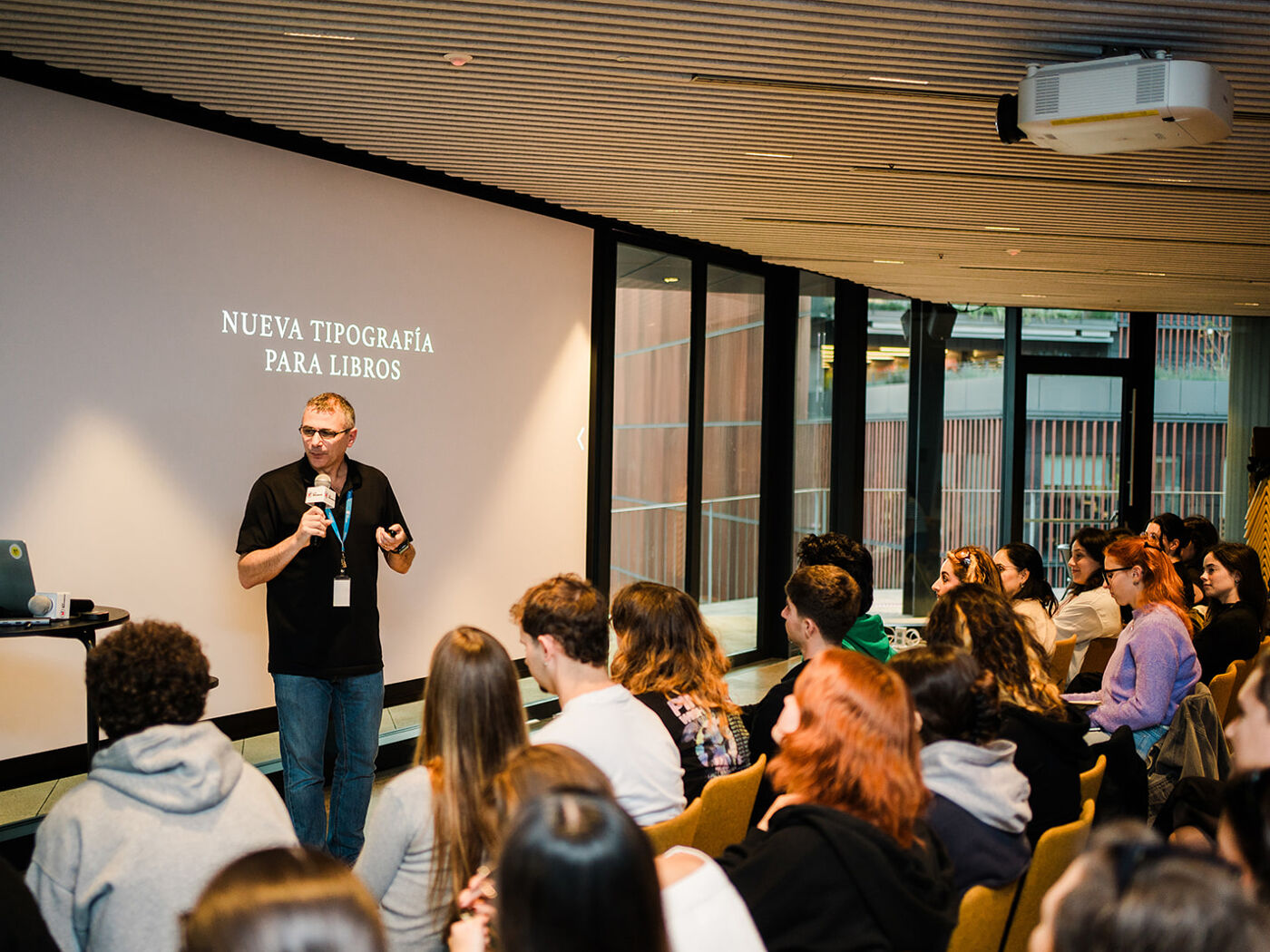 A presenter is addressing an audience in a conference room, with the presentation title "NUEVA TIPOGRAFÍA PARA LIBROS" displayed on a large screen behind him.