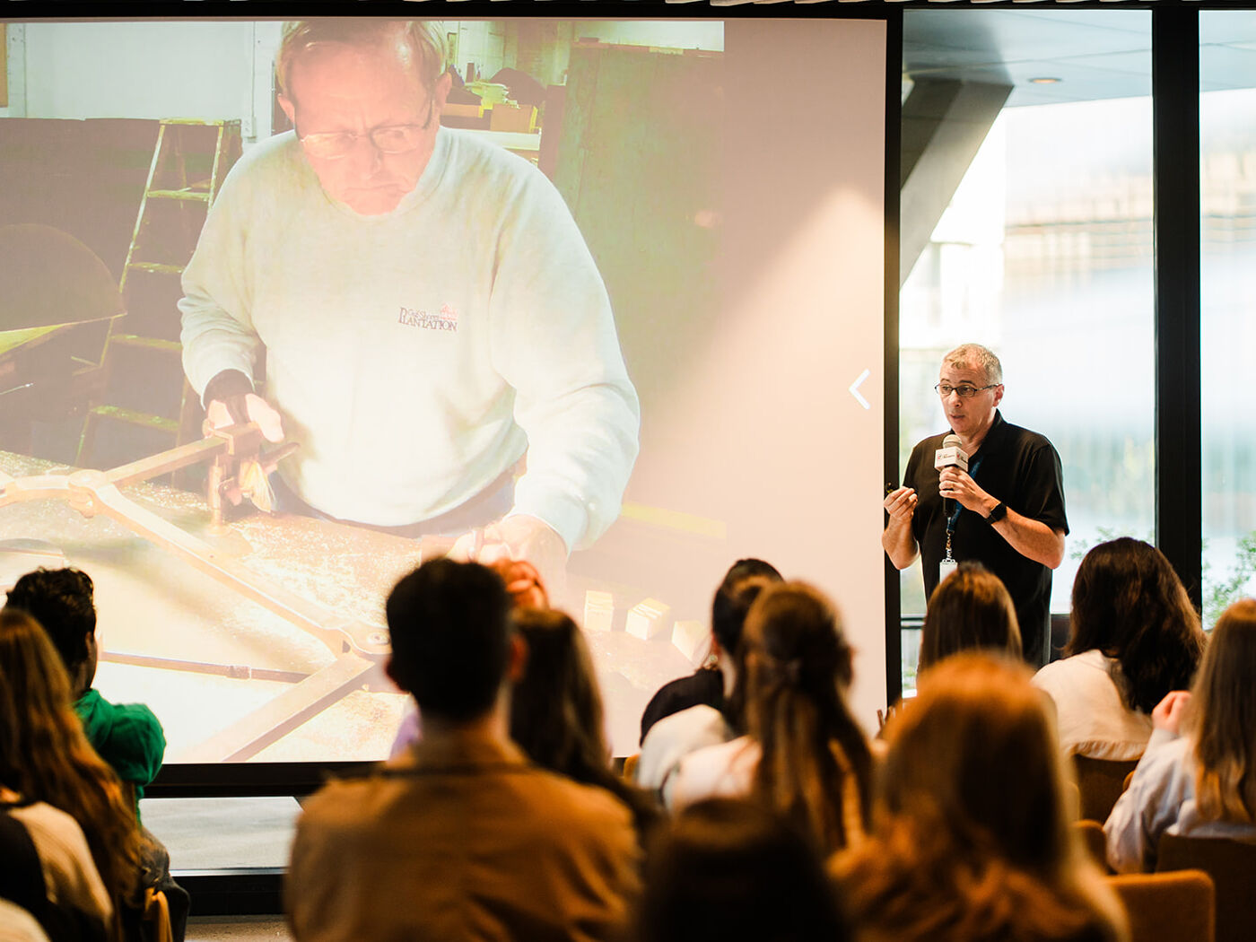 A man is giving a presentation to an audience, with a large screen behind him displaying a close-up of another man working on a craft.