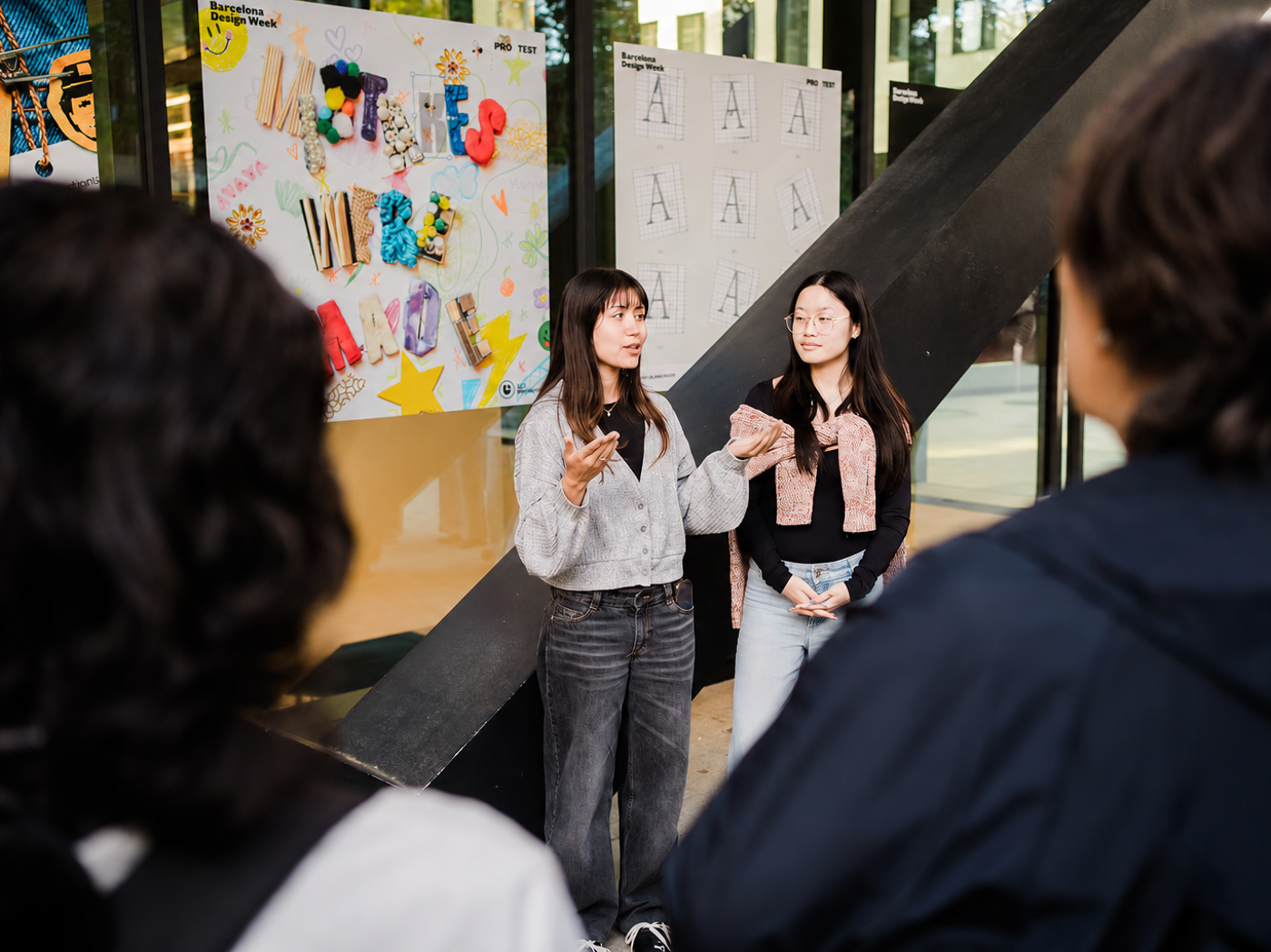 Two young women are presenting a project, possibly art or design related, to a small group of people. The presentation appears to be outdoors, with posters and displays visible in the background.