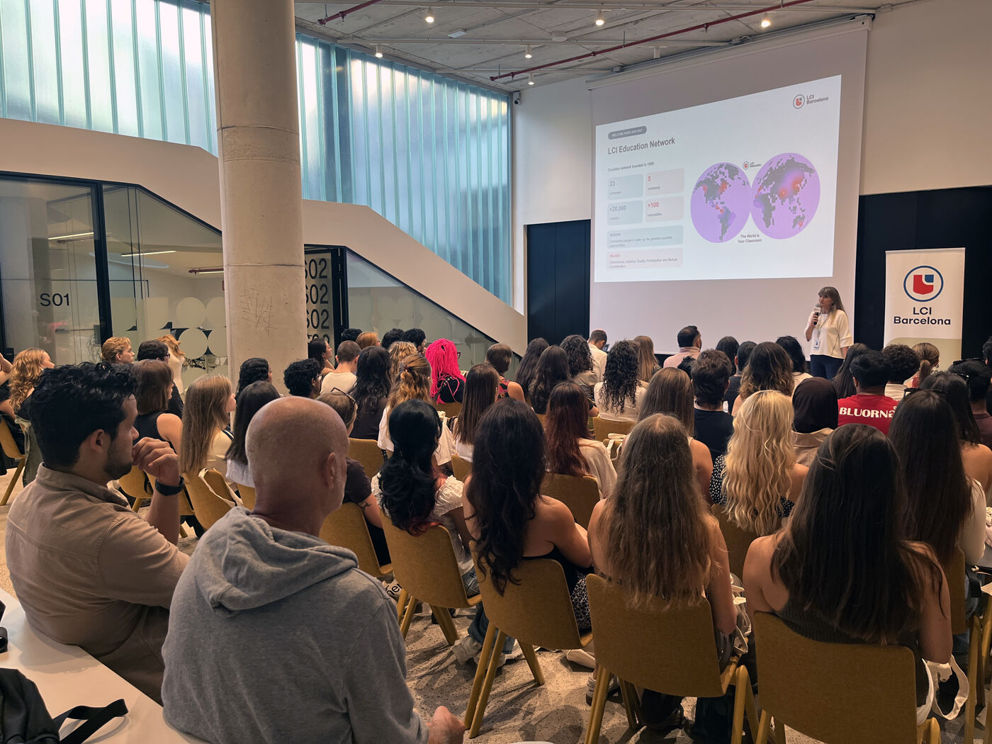 The image shows a presentation or lecture taking place in a modern, well-lit room with a large screen at the front. An audience is seated, facing the presenter.
