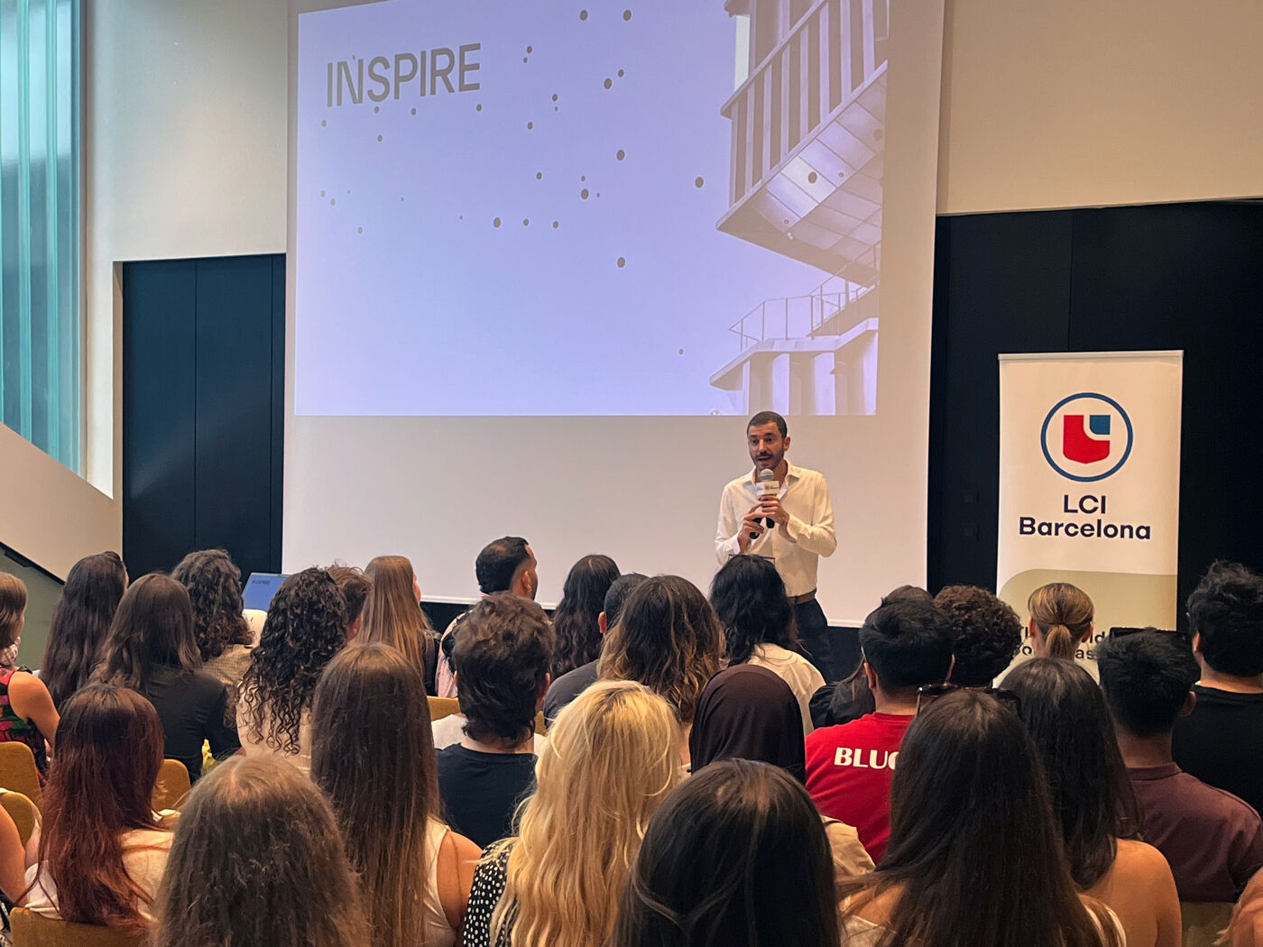 A speaker presents the word 'INSPIRE' on a screen to an audience seated in a lecture hall. A banner for LCI Barcelona is visible on the right.