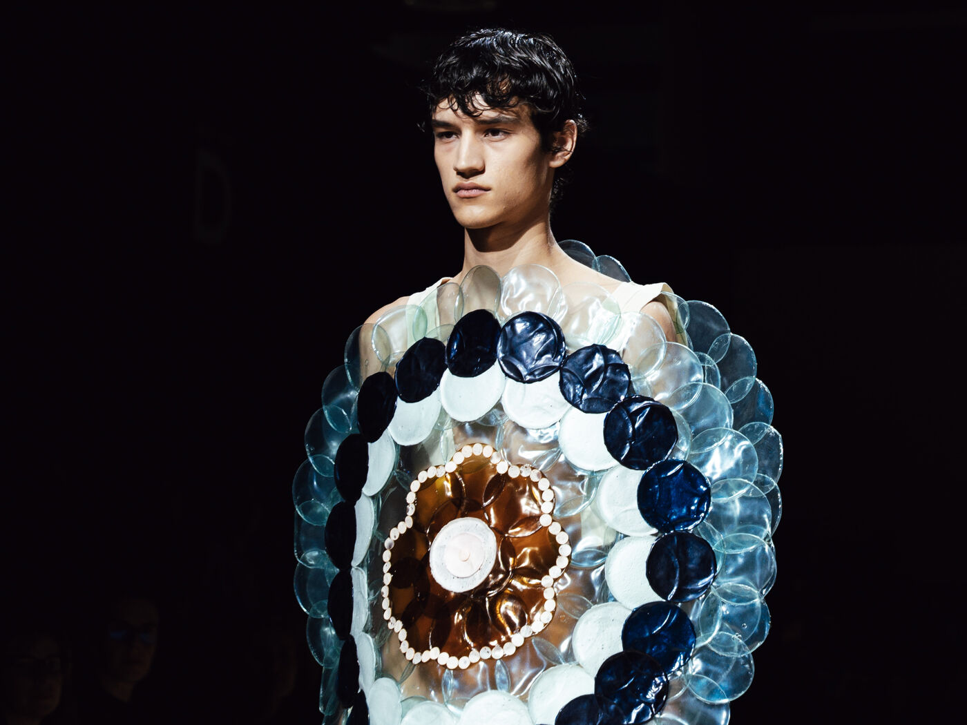 A male model showcases a unique garment featuring a circular, flower-like design. Crafted from translucent and opaque materials, it evokes the appearance of a jellyfish or a flat disc.