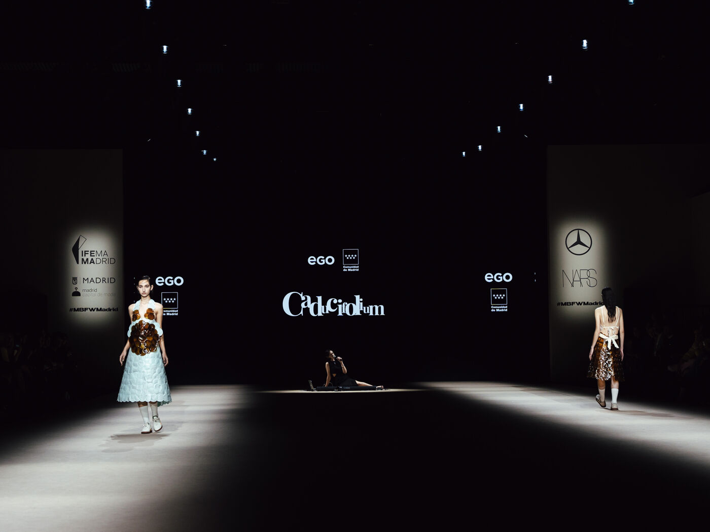 Models walk the runway at a fashion show, showcasing unique designs against a dark backdrop with illuminated branding and stage lighting.