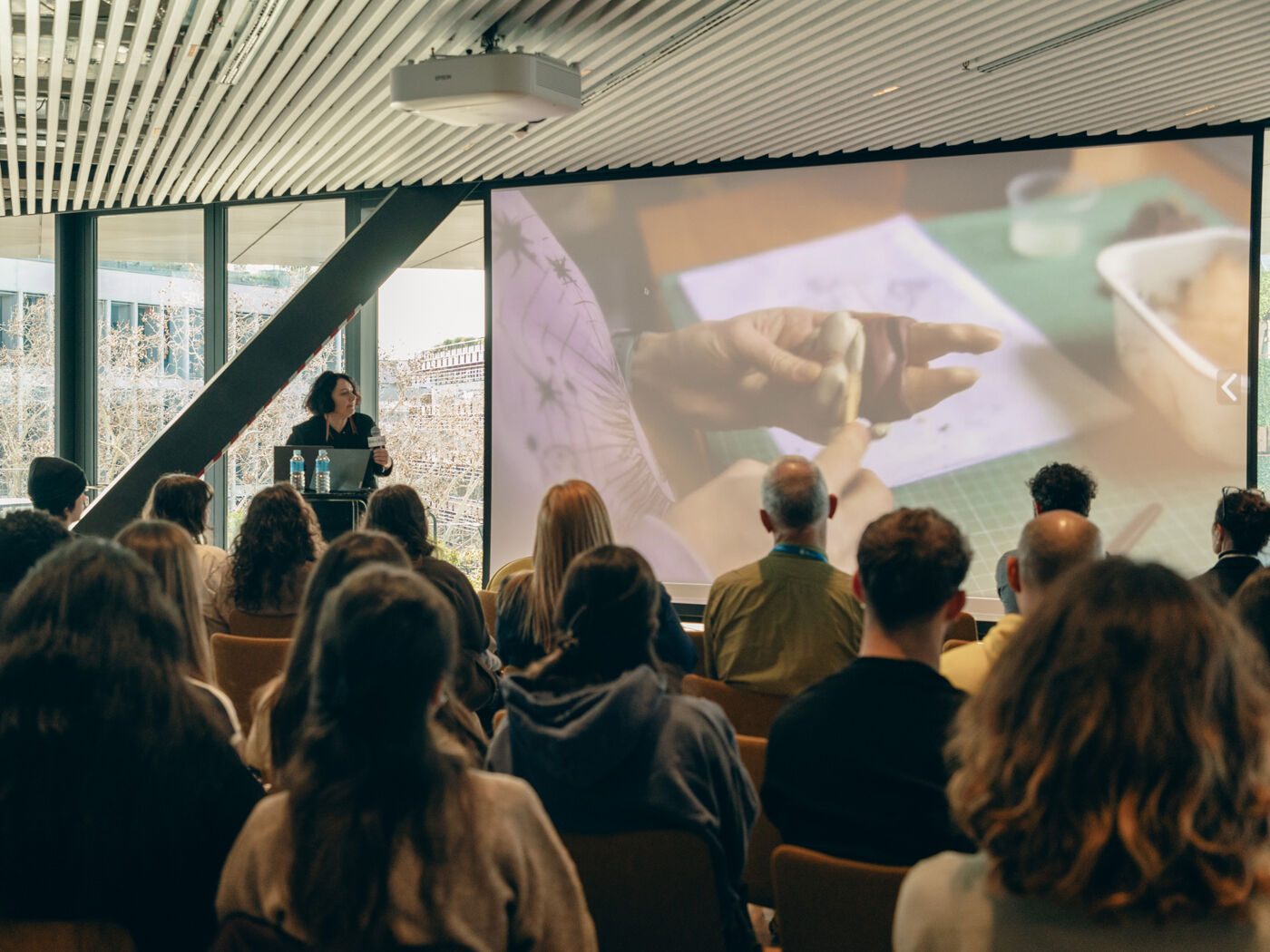 A woman presents to a large audience while an image is projected on the large screen behind her, possibly a lecture or presentation.