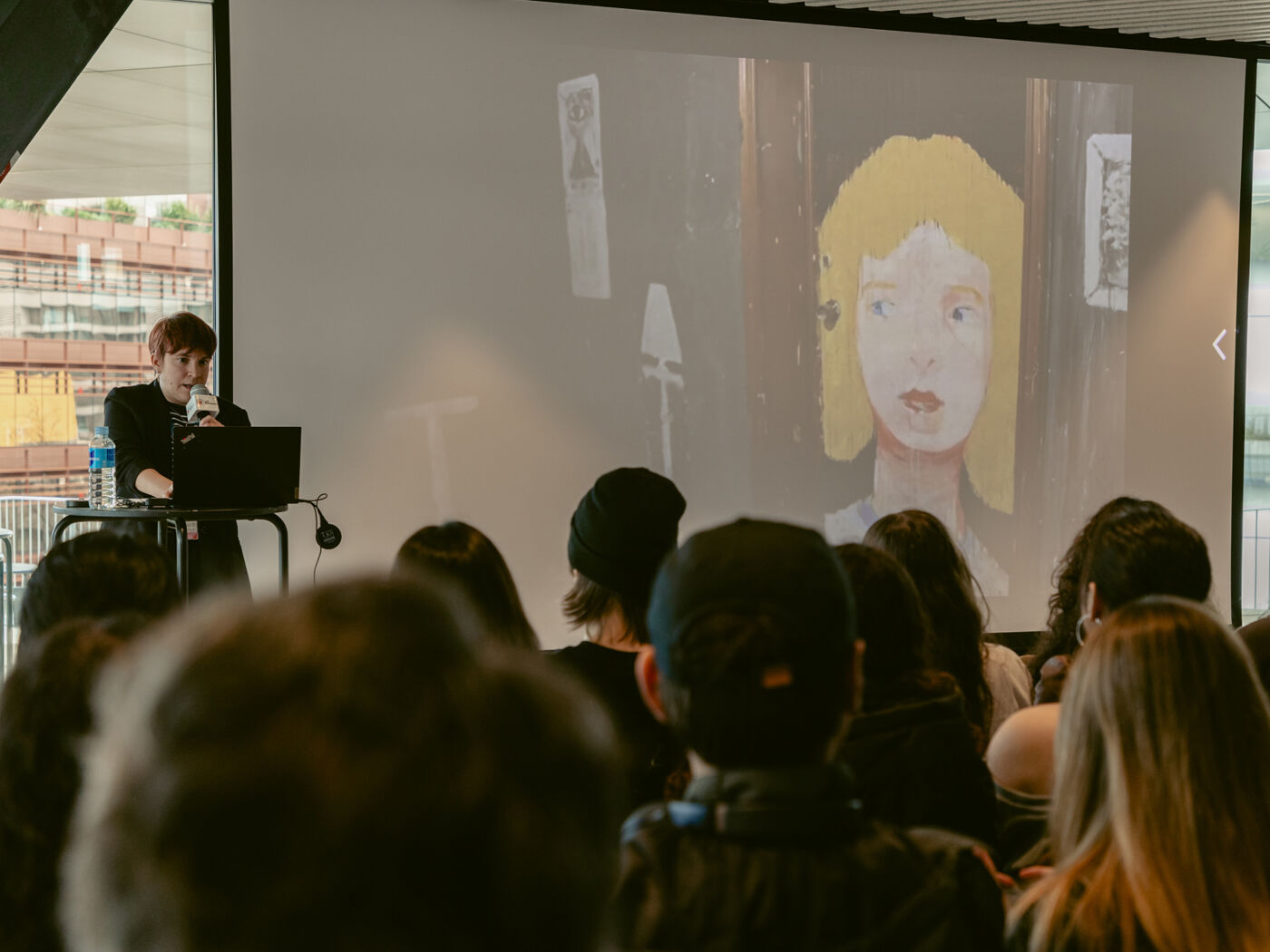 A person is presenting in front of a large screen displaying a portrait of a woman with blonde hair and large eyes. The presentation appears to be taking place in a casual setting with an audience seated.