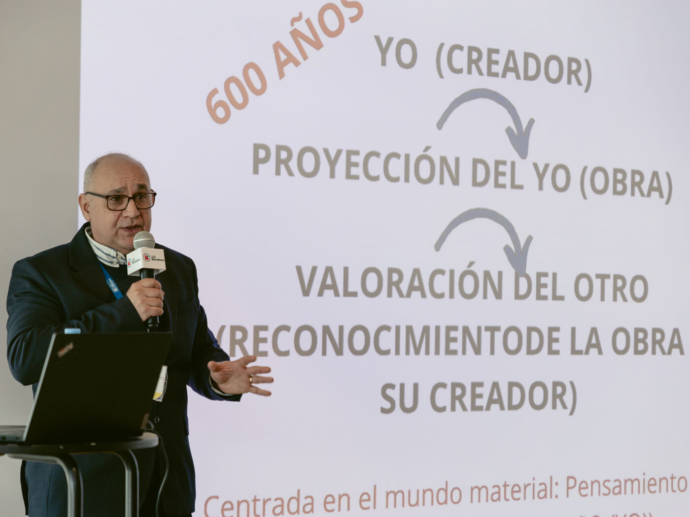 A man speaks in front of a screen displaying a diagram about the creative process, including the creator, the work, and its valuation.