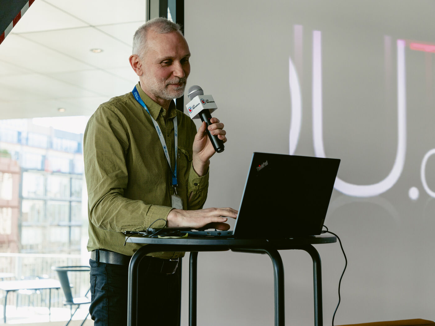 A middle-aged man with gray stubble on his chin speaks into a microphone while working on a laptop in front of a large screen.