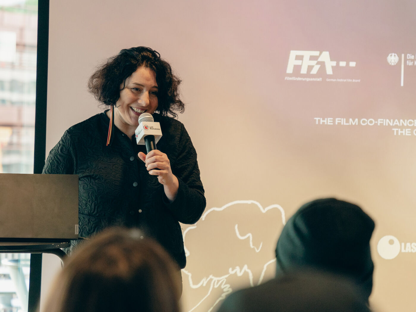 A woman smiles while speaking into a microphone in front of an audience, with a laptop beside her and a backdrop featuring logos.