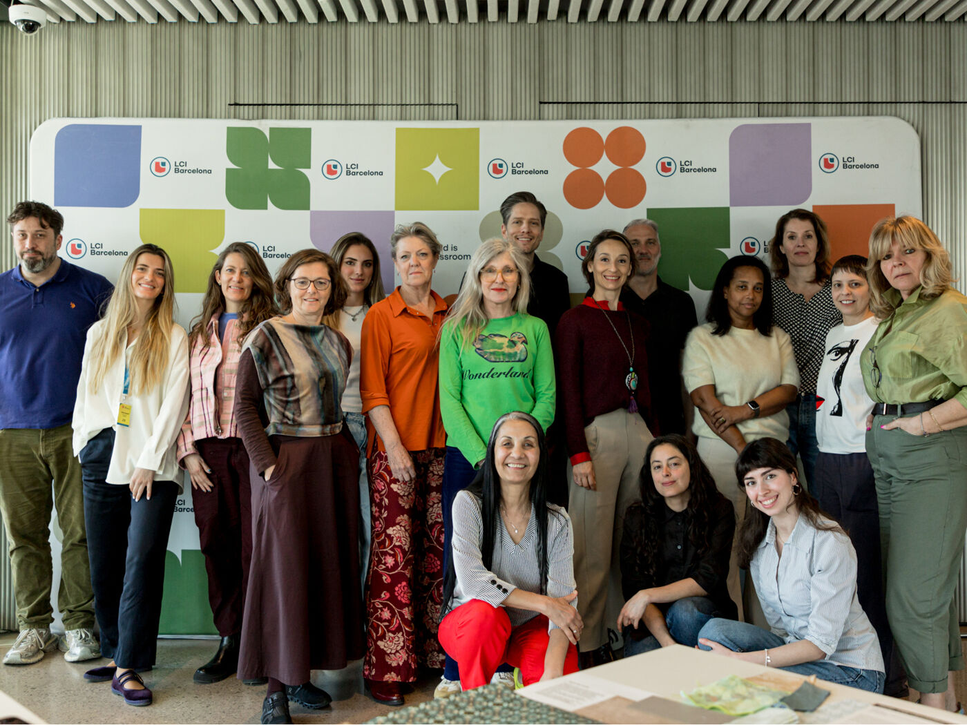 A diverse group of men and women are posing together indoors in front of a large backdrop featuring a colorful logo. They appear to be at a gathering or event.