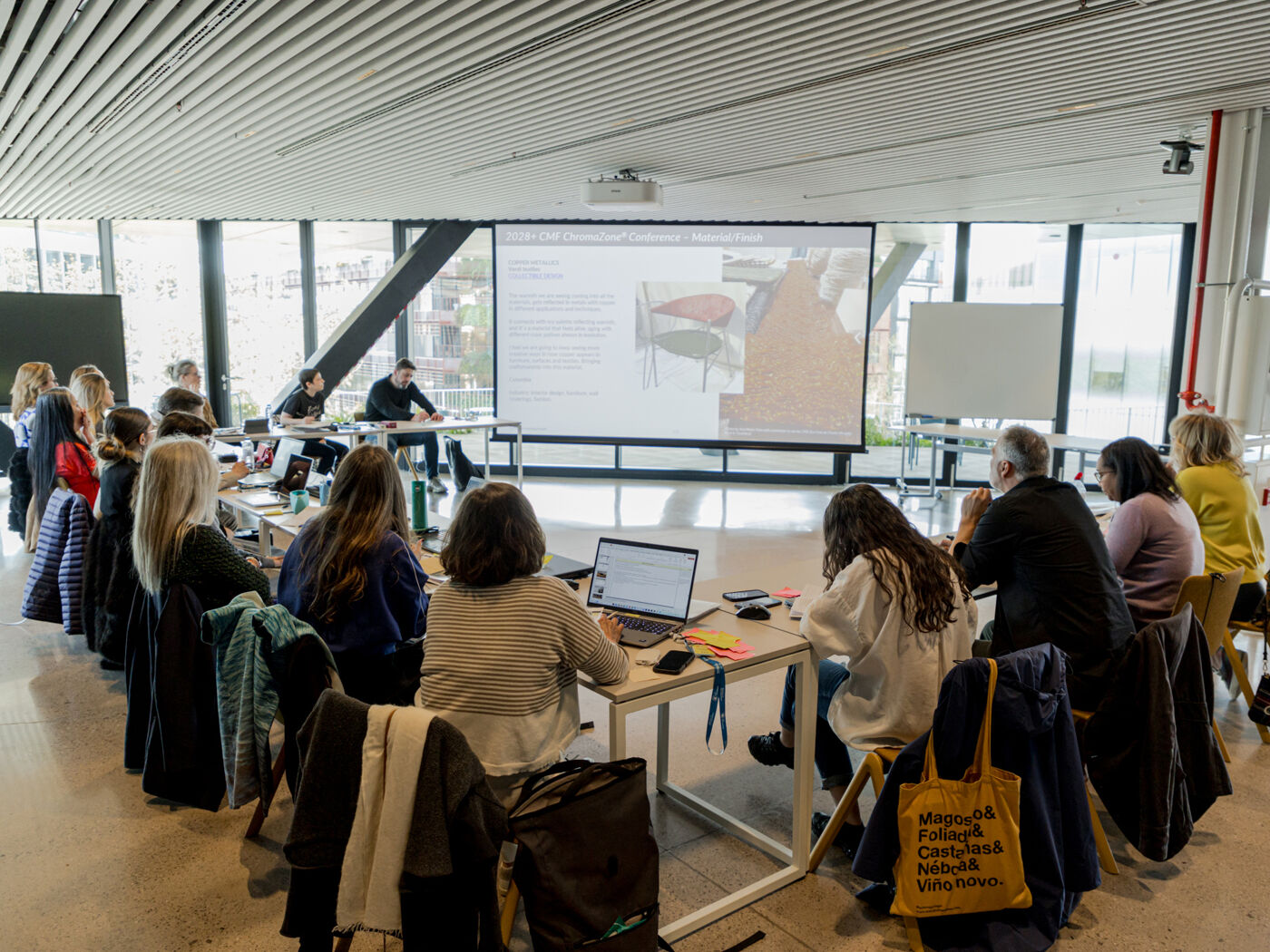 A group of people are attending a presentation or workshop, with a large screen in the background displaying images and a speaker presenting.