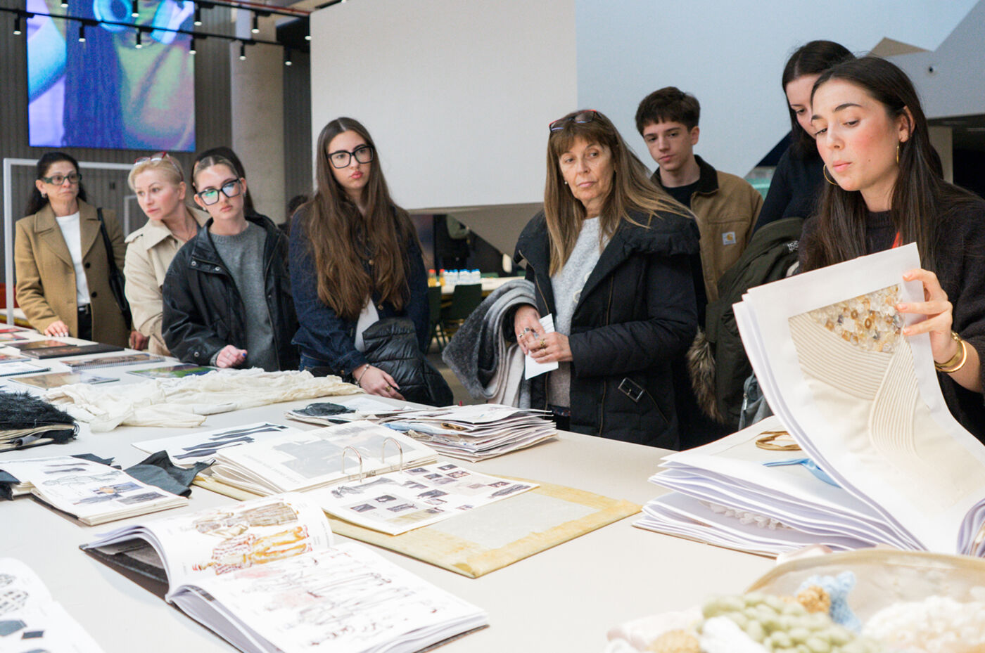 A group of people are gathered around a table, examining papers and designs. They appear to be discussing or reviewing the displayed materials, possibly in a design or art context.