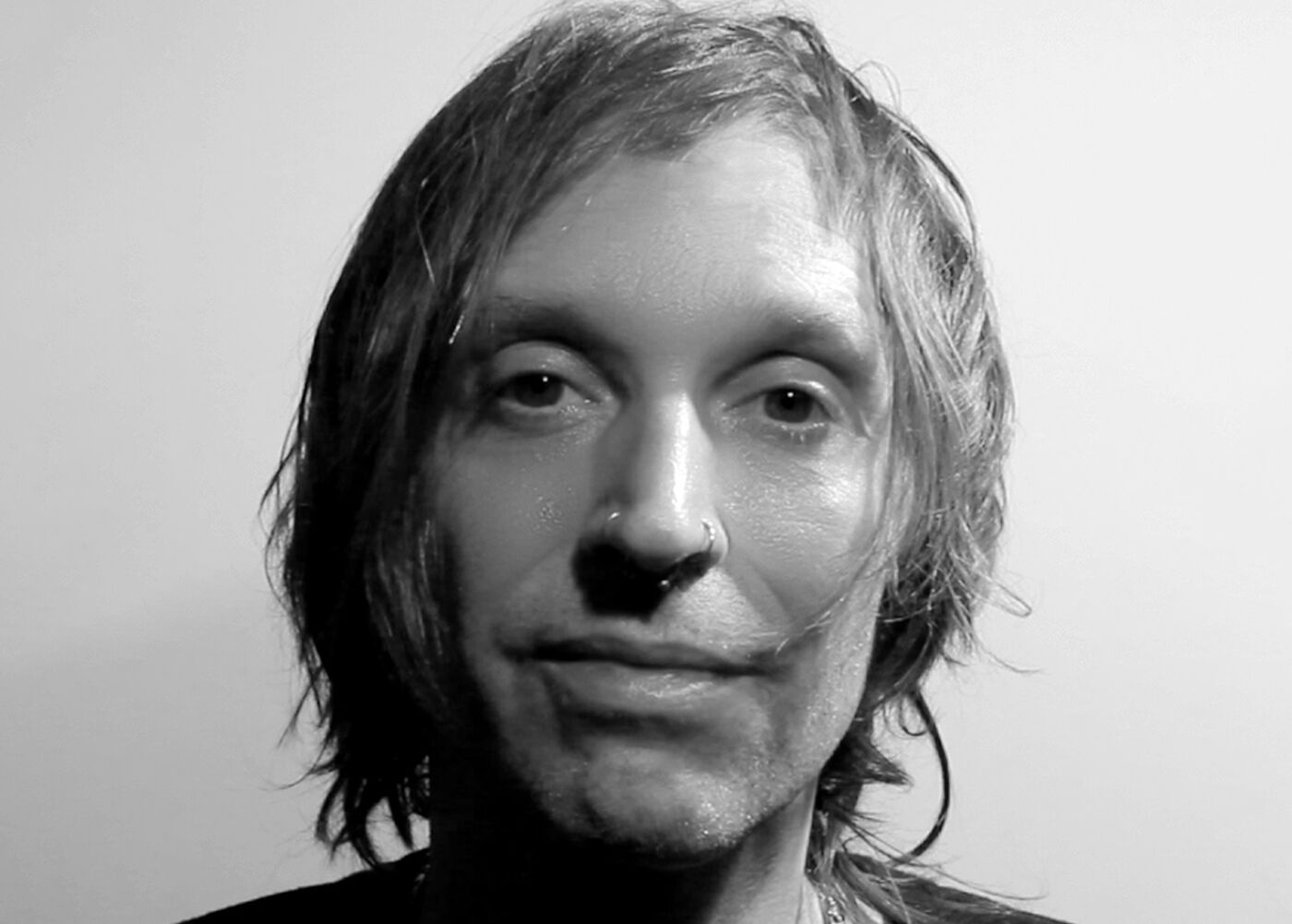 A close-up black and white portrait shows a man with shoulder-length hair and a nose ring, looking directly at the camera with a neutral expression.