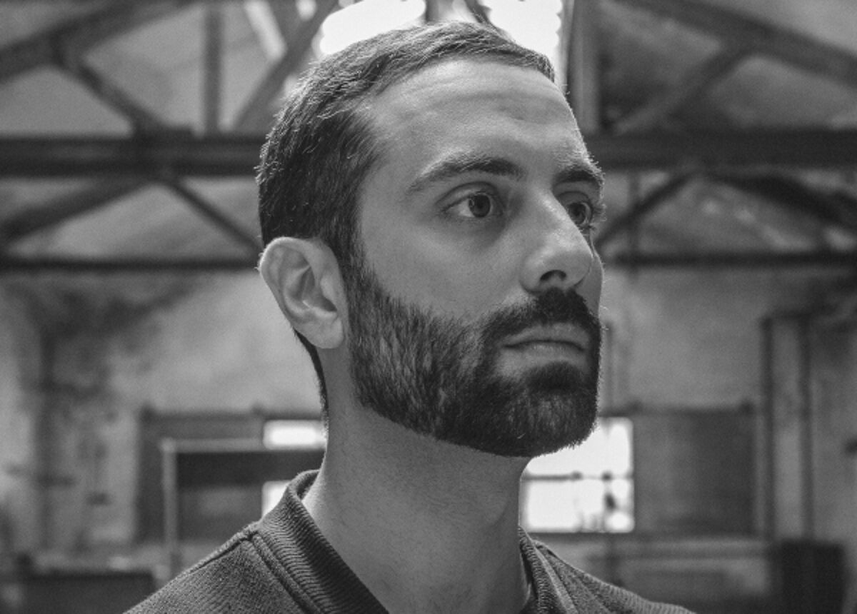 Close-up black and white shot of a man with a beard, looking to the right, in what seems to be an industrial setting.