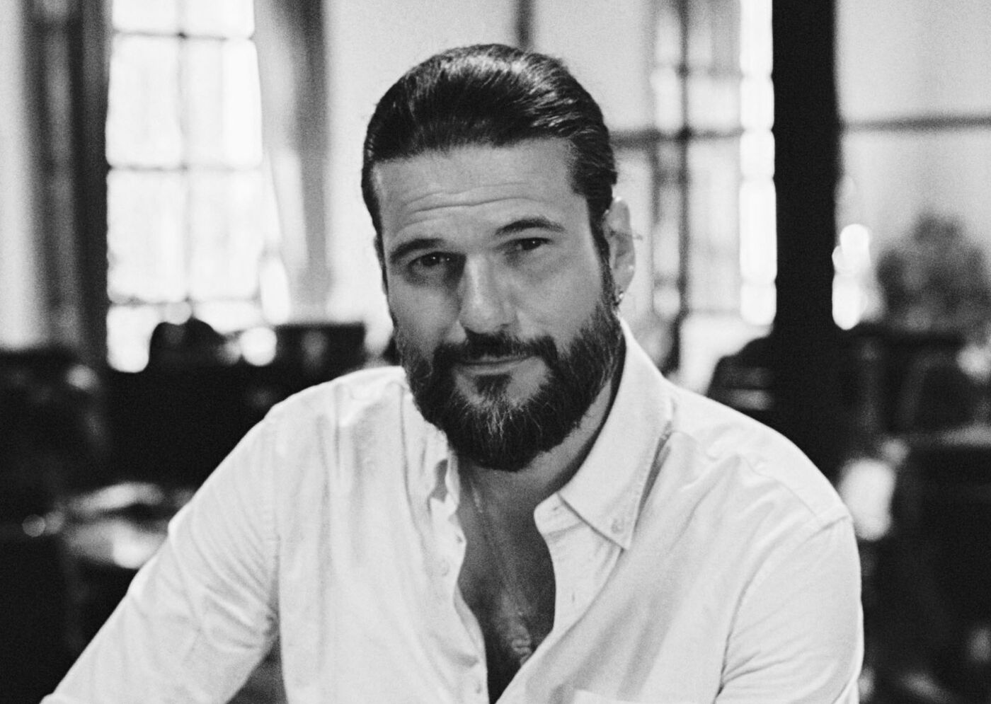 This is a black and white portrait of a man with a beard, wearing a white collared shirt. He has dark hair slicked back and is looking directly at the camera with a serious expression.
