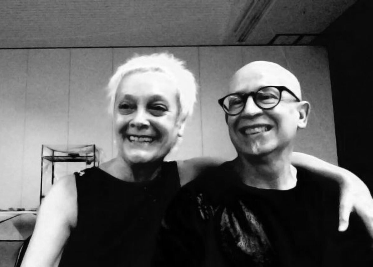The image shows a black and white photo of a smiling woman with short white hair and a smiling bald man wearing glasses. They are standing close together with the man's arm around the woman's shoulder.