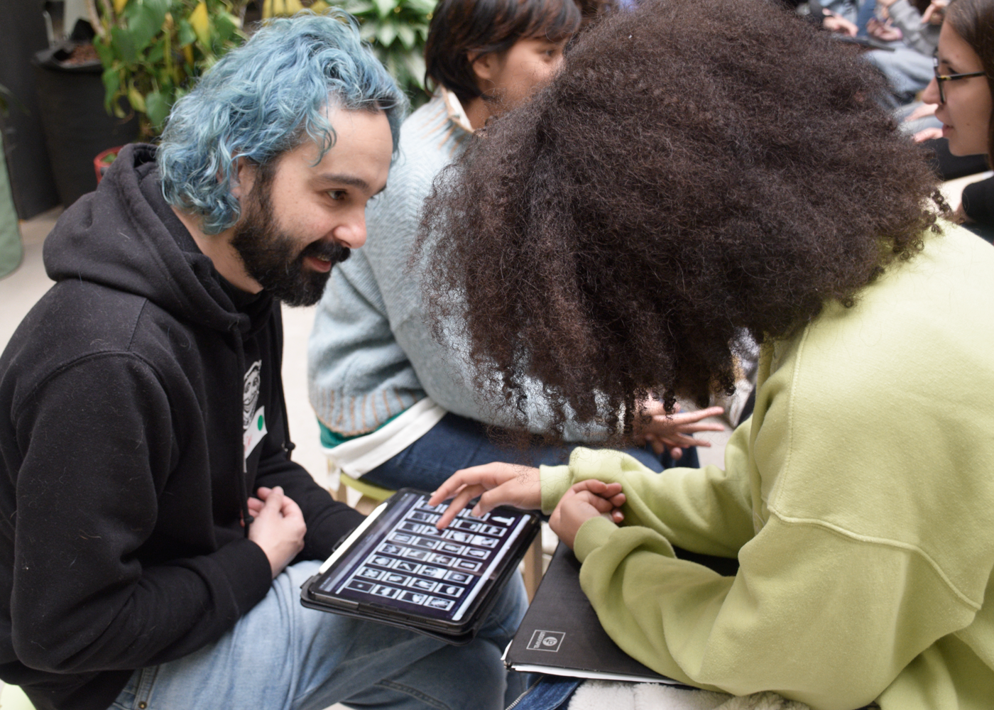 Two people engage in a lively discussion over a tablet, surrounded by others in a workshop setting.