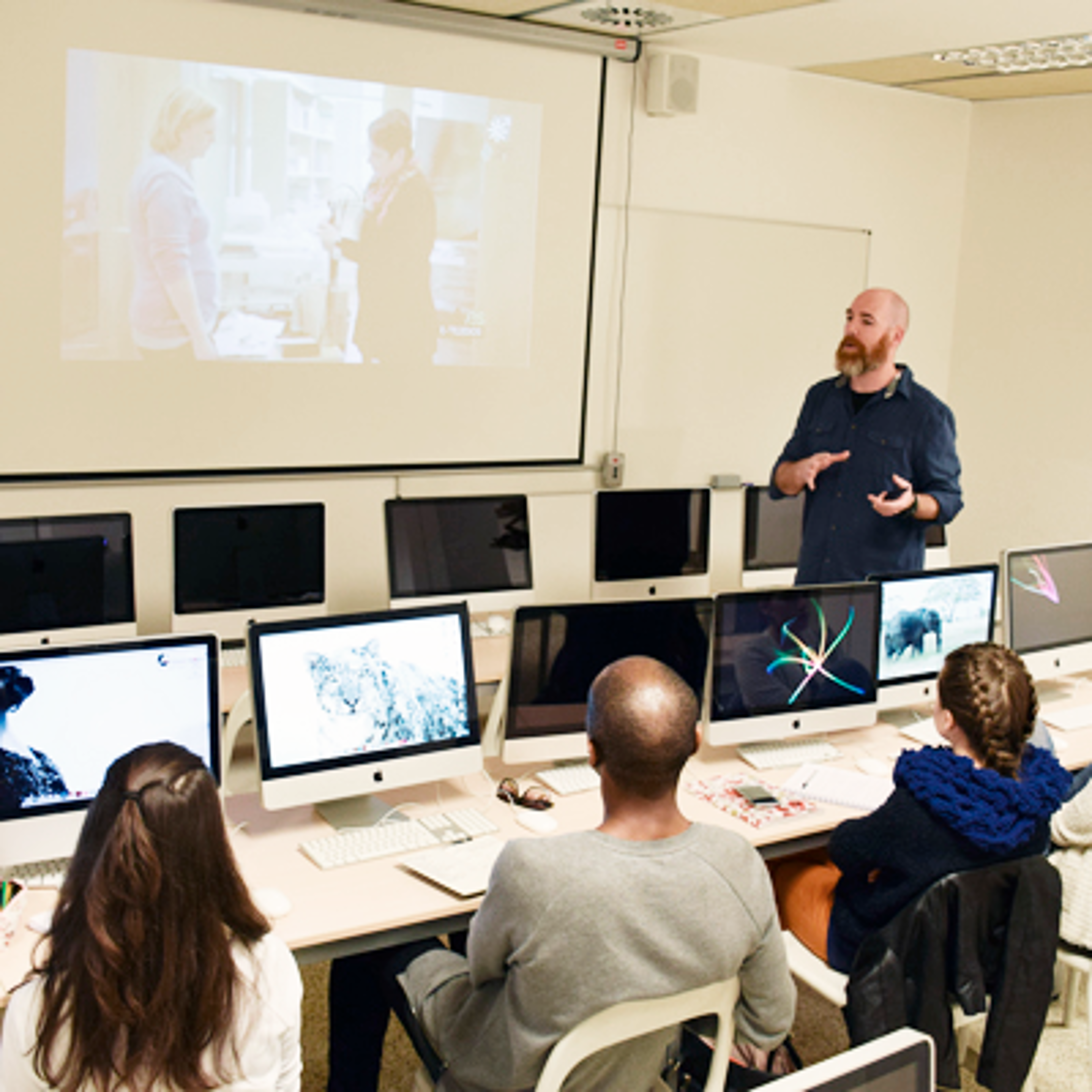 Profesor enseñando diseño gráfico a estudiantes trabajando en iMacs, con una pantalla de presentación al fondo.