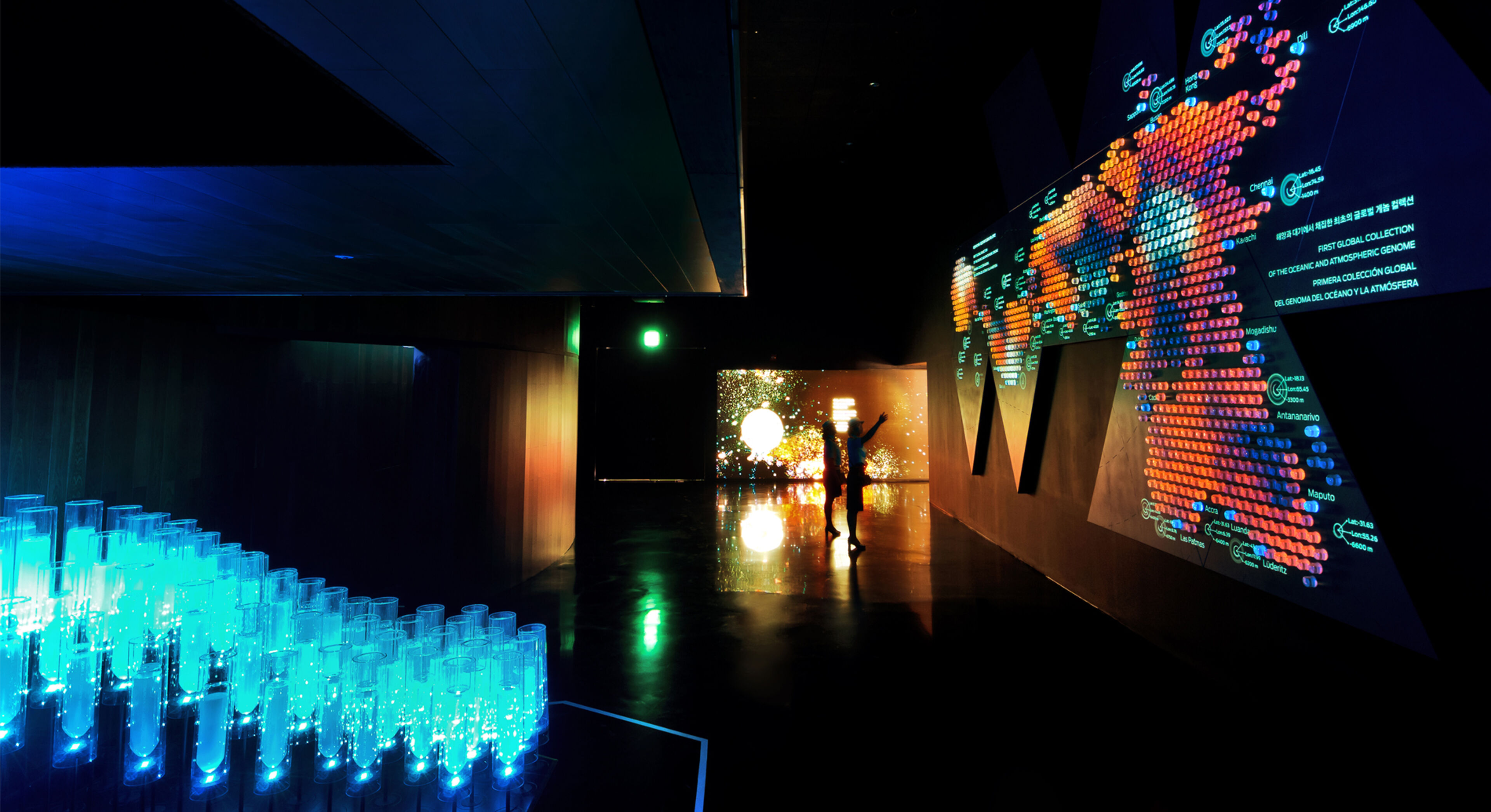 Una habitación oscura iluminada por una instalación de luz interactiva con pantallas visuales vibrantes y la silueta de una persona interactuando con la exhibición.