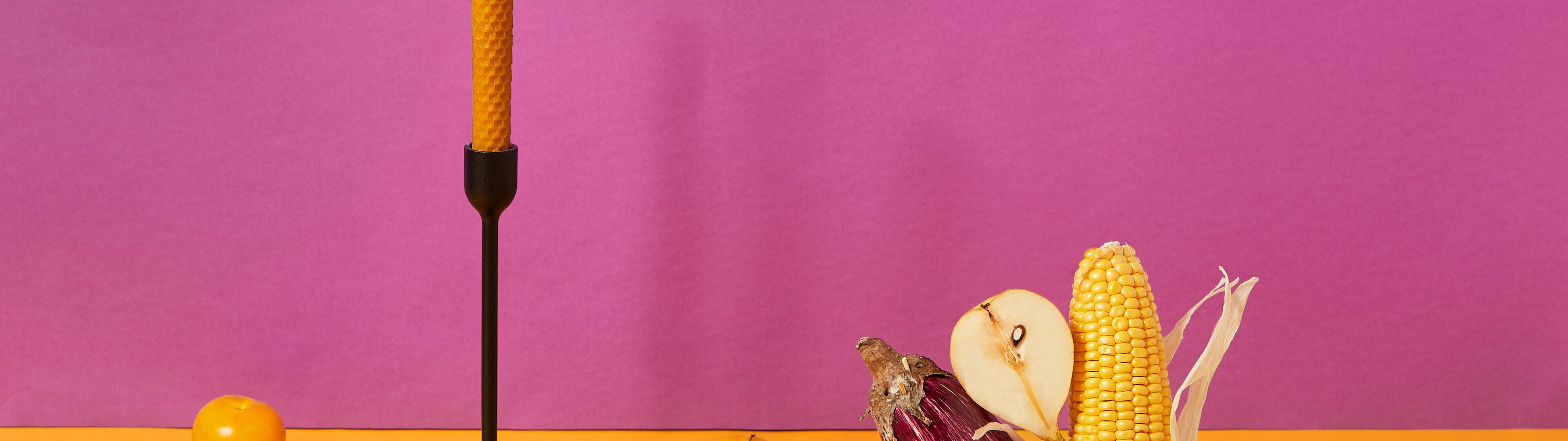 Una composición de bodegón colorida con una vela iluminada, frutas frescas y verduras sobre un fondo de dos tonos.