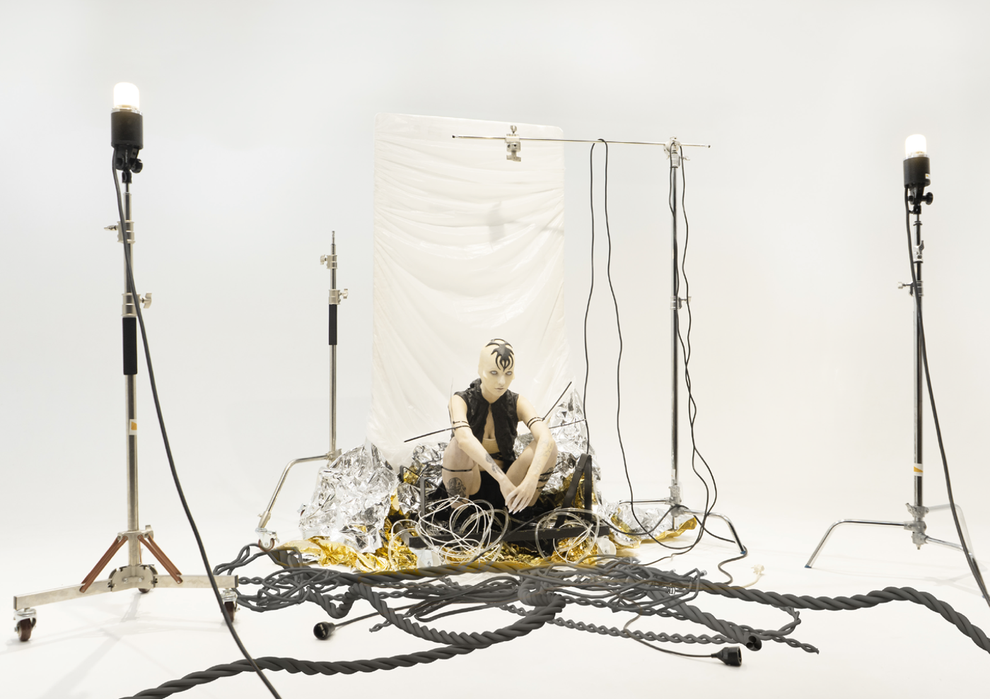 A minimalist art installation featuring a model seated amidst tangled cables, metallic foil, and industrial lighting. The stark white background contrasts with the intricate setup, evoking themes of technology, human connection, and modernity.