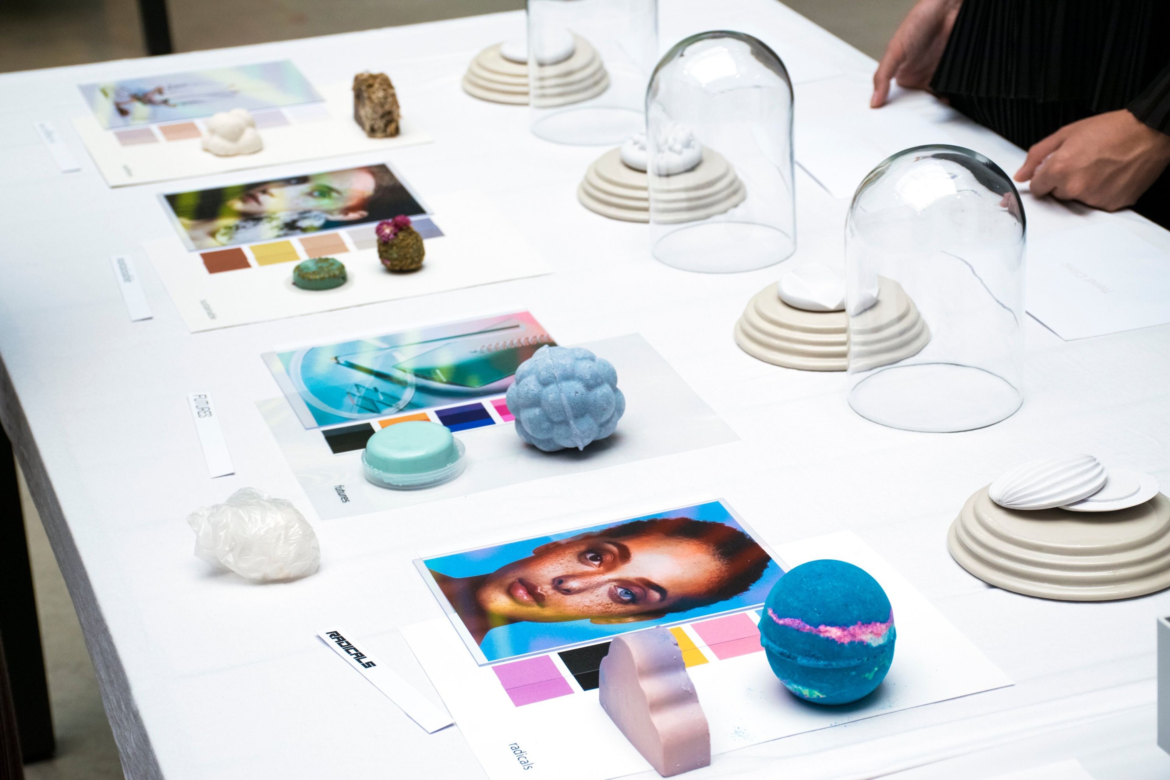 Una mesa que muestra varios elementos de diseño, incluyendo fotografías, muestras de color y modelos impresos en 3D bajo cúpulas de vidrio."