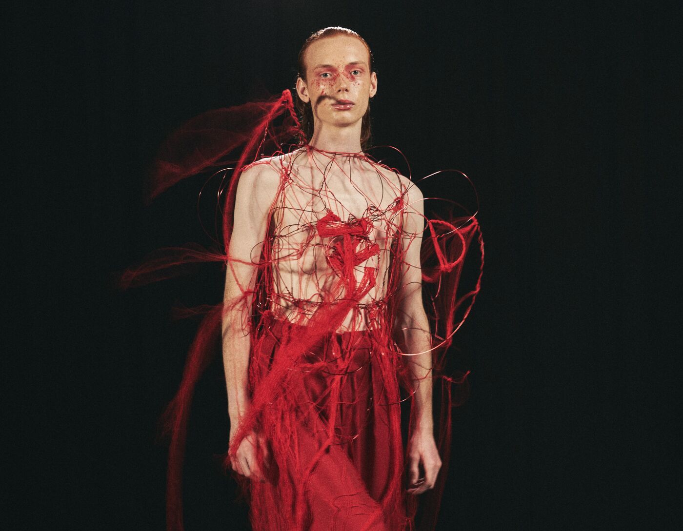 A model wearing an avant-garde fashion piece featuring intricate red threads and abstract patterns. The outfit combines bold textures with a deconstructed aesthetic, blending art and couture. The backdrop is dark, emphasizing the vibrant design.