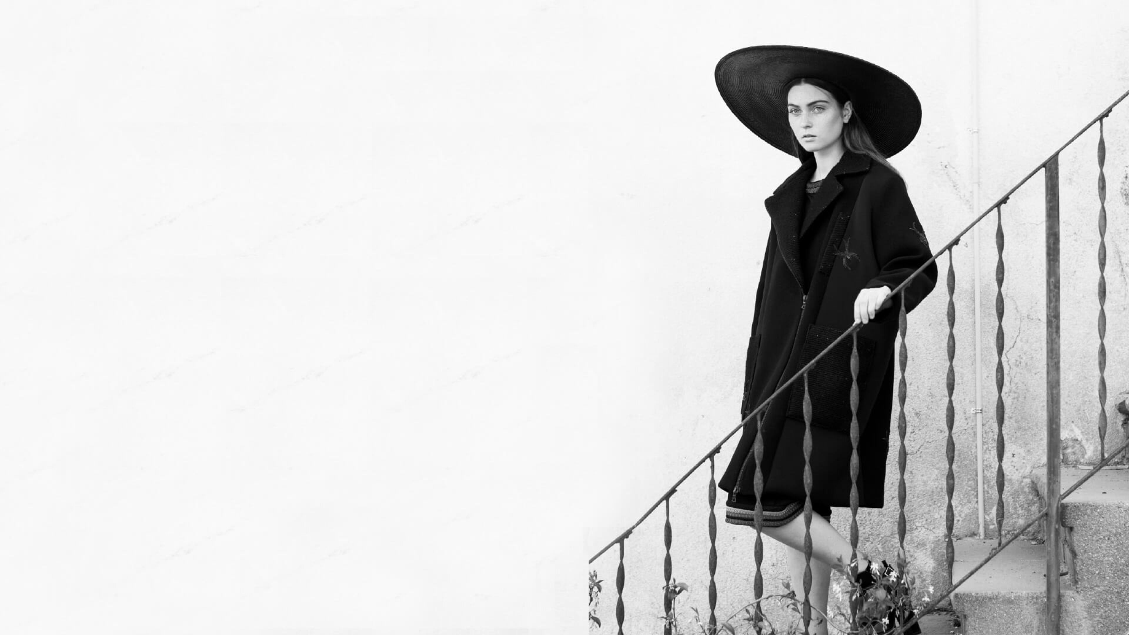 Un model amb abric negre i barret de volta ampla posa en una escala exterior, evocant un estil monocromàtic atemporal.